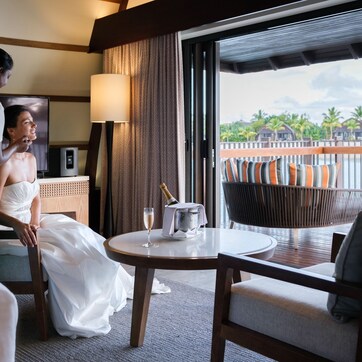 A hotel room with a bed, a table with a bottle of champagne, a chair, and a view of palm trees through large windows.