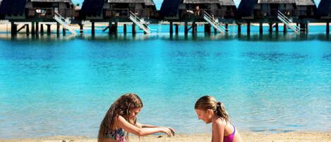 Two people playing with sand on a beach with overwater bungalows in the background.