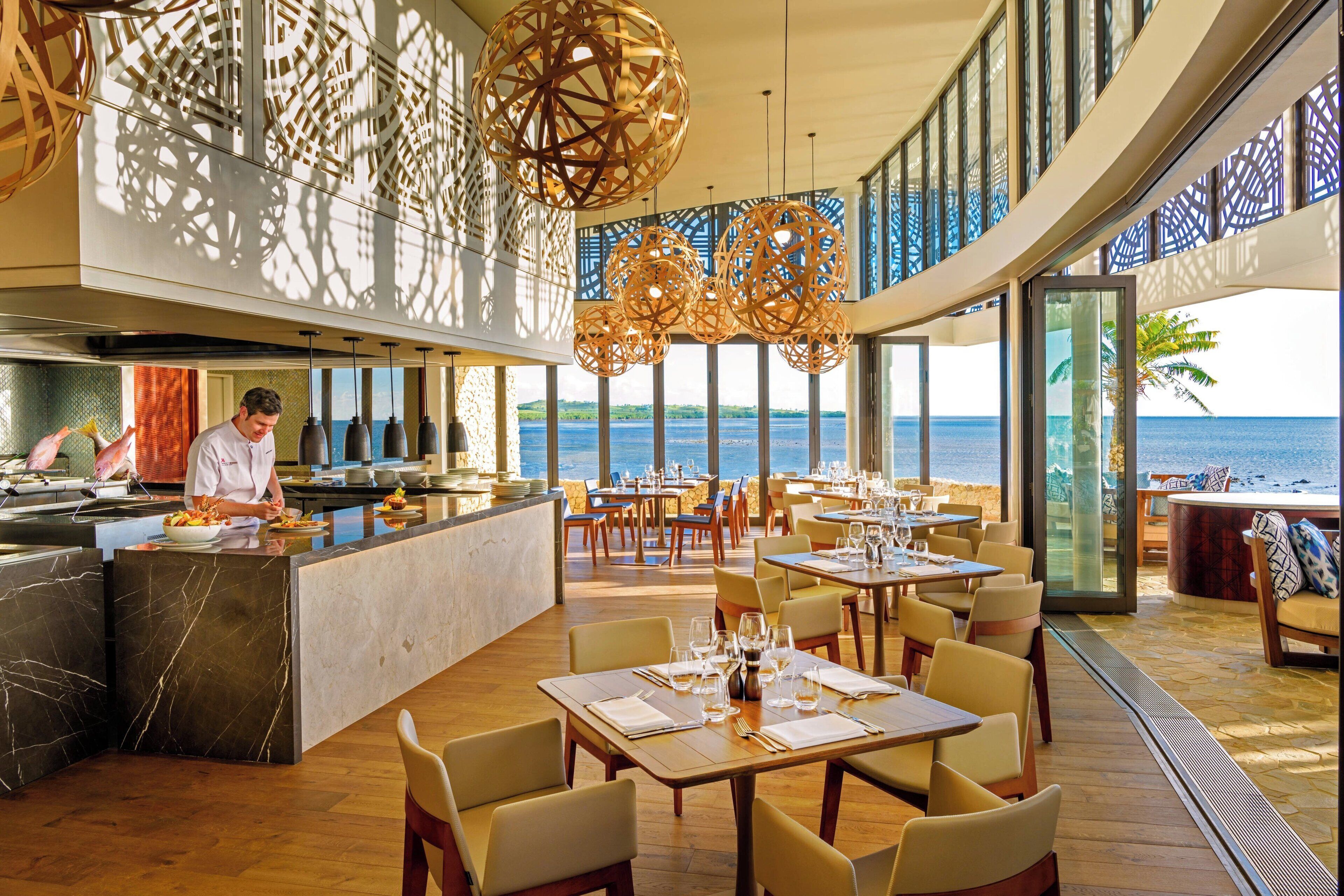 A restaurant with a view of the sea, featuring a chef preparing food, wooden tables, and a modern interior design.