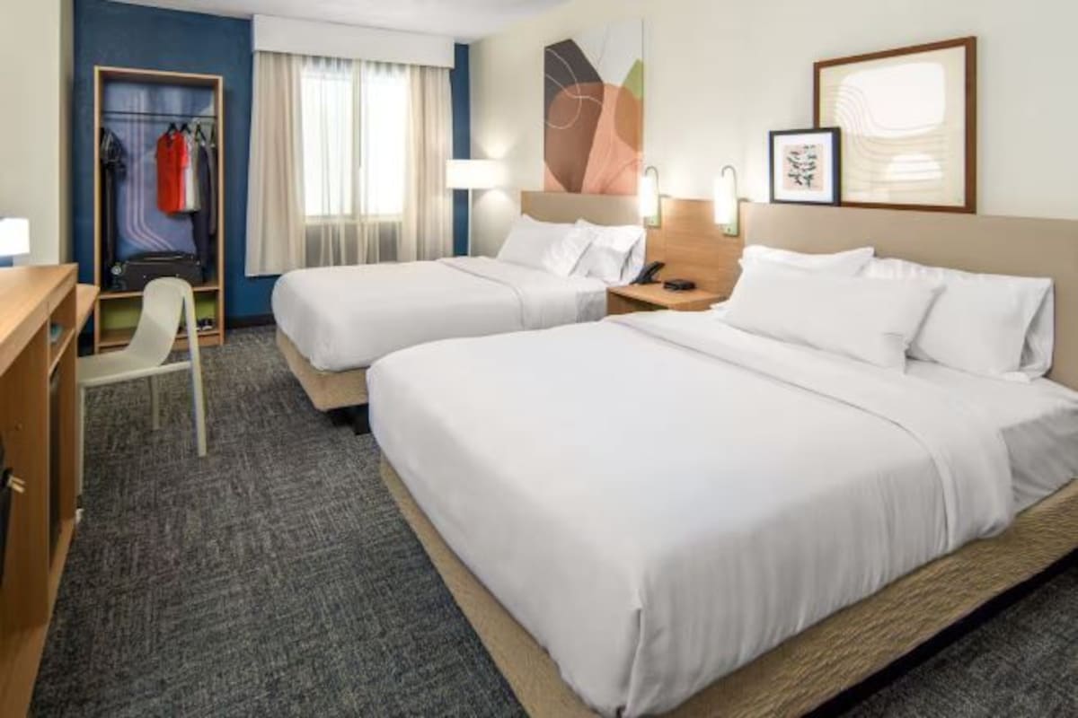 A hotel room with two single beds, a desk, a chair, a wardrobe, and a large abstract painting on the wall.