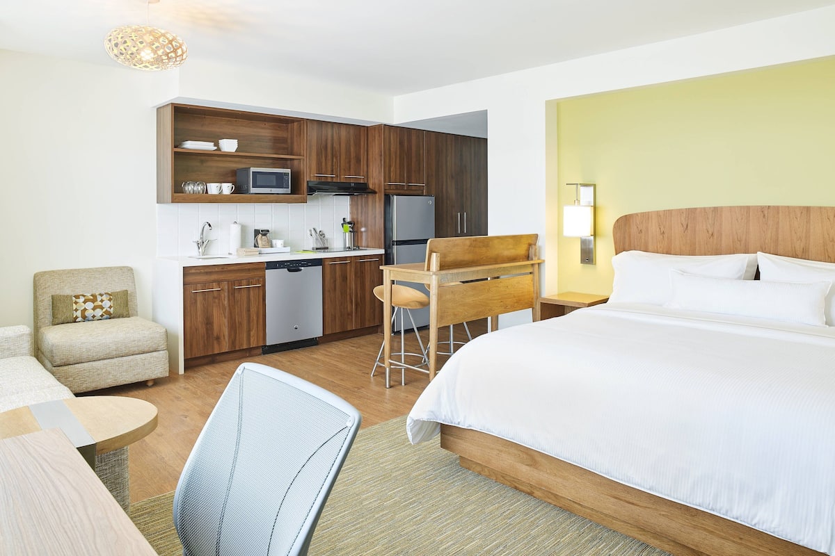 A hotel room with a bed, a desk, a chair, a kitchenette, and a dining area.