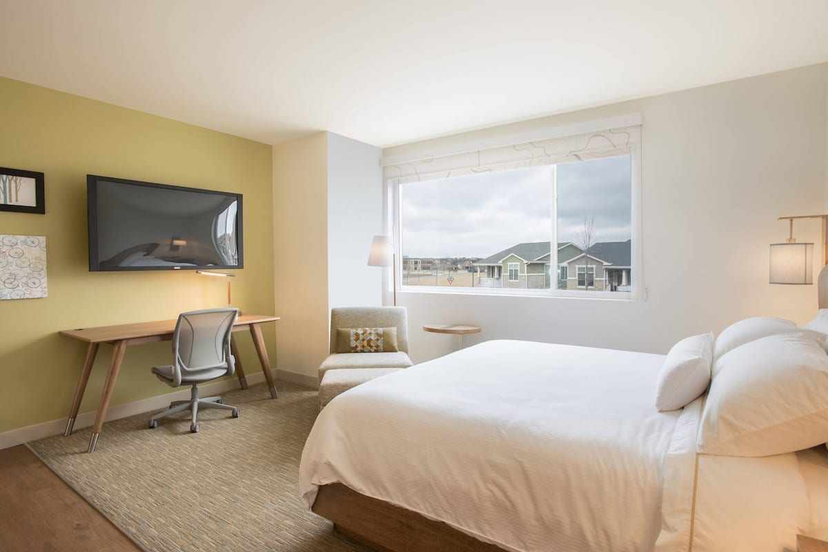 A hotel room with a bed, desk, chair, TV, and a window with a view of houses.