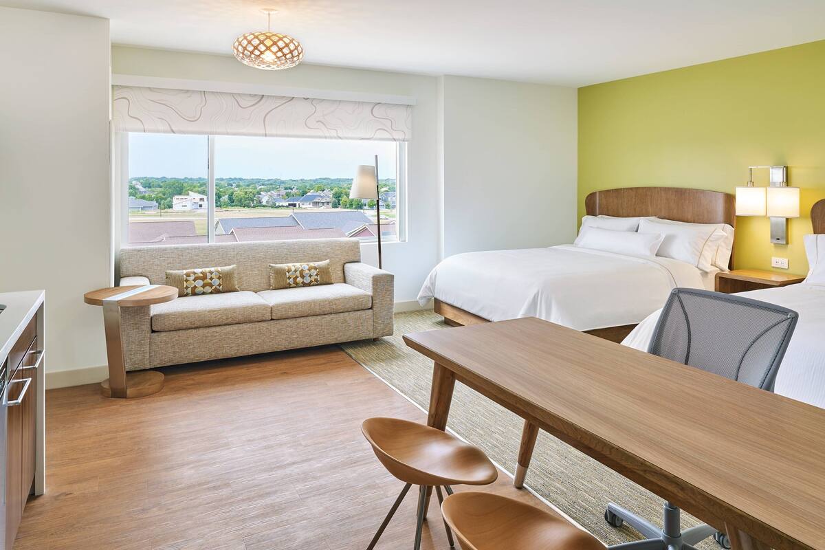 A hotel room with a bed, a sofa, a dining table, and a view of the outside.