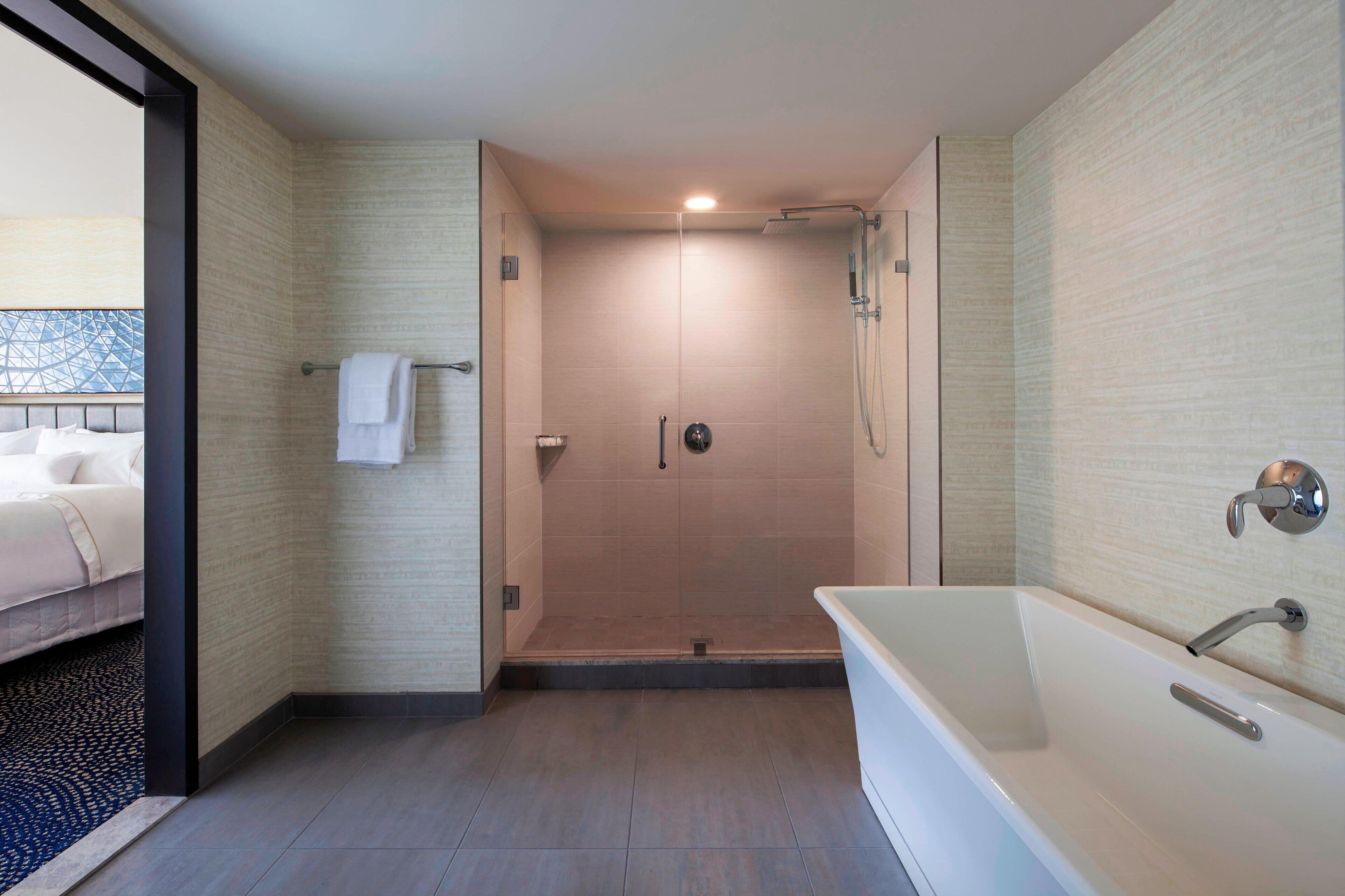 A modern bathroom with a walk-in shower, a bathtub, and a visible bed with a towel.