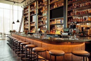 A bar area with a long counter, multiple stools, and shelves stocked with bottles. - The Westin Milwaukee (Milwaukee)