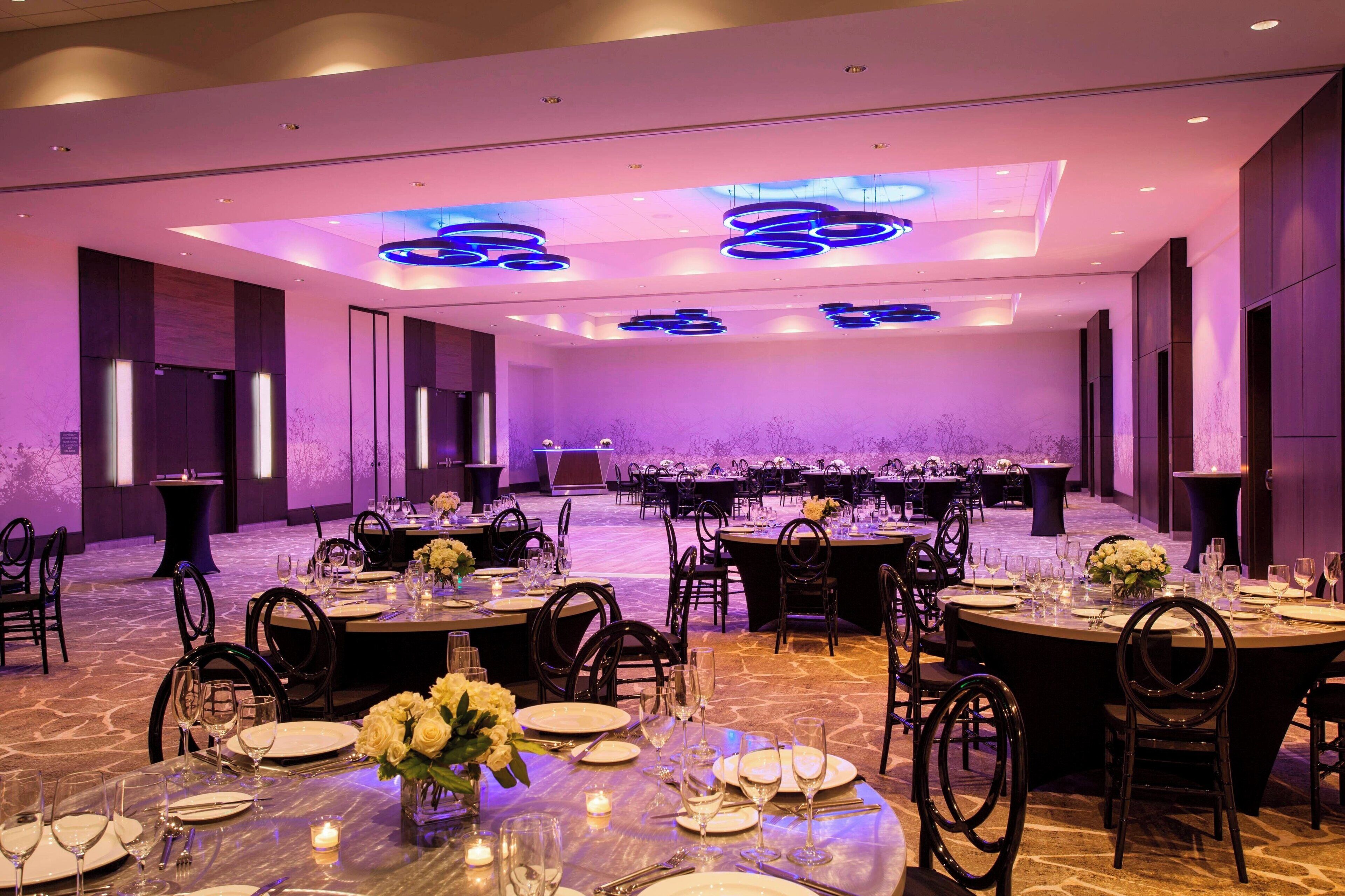 Salão de banquetes com mesas redondas preparadas para refeições, equipado com cadeiras, pratos, copos e velas.