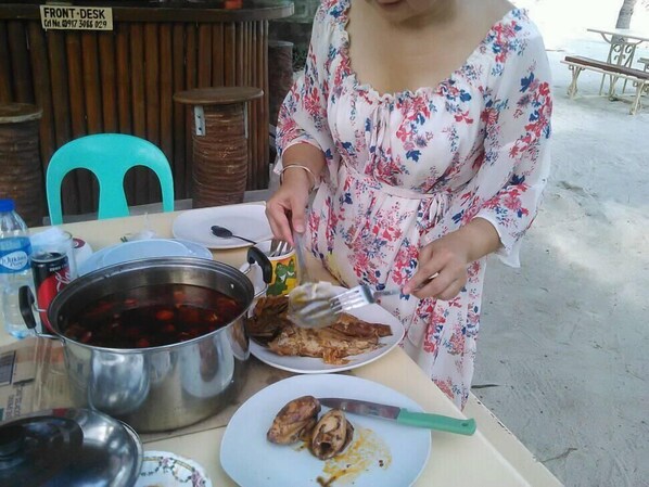 Una persona sirve comida en una mesa al aire libre, con una olla de guiso, platos de comida y una botella de agua.