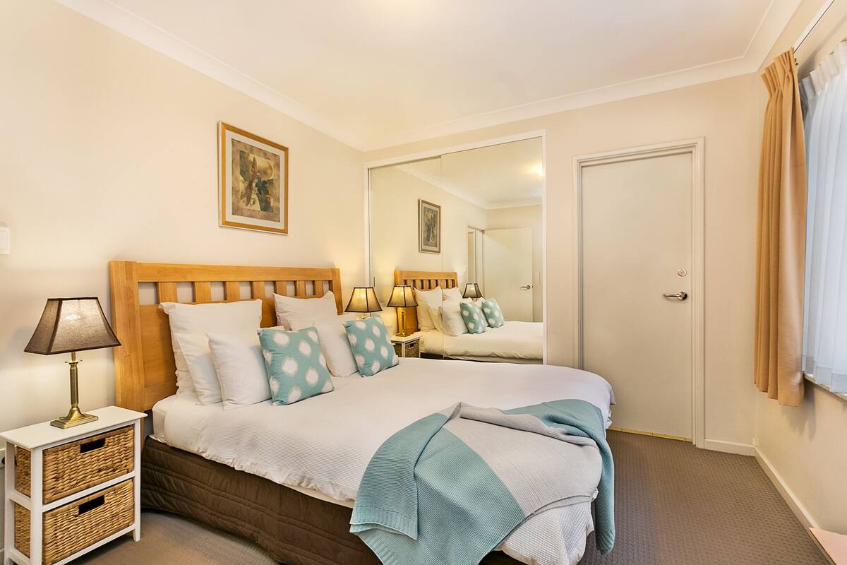 A neatly arranged bedroom with a wooden headboard, a large mirror, and a bedside table with lamps.