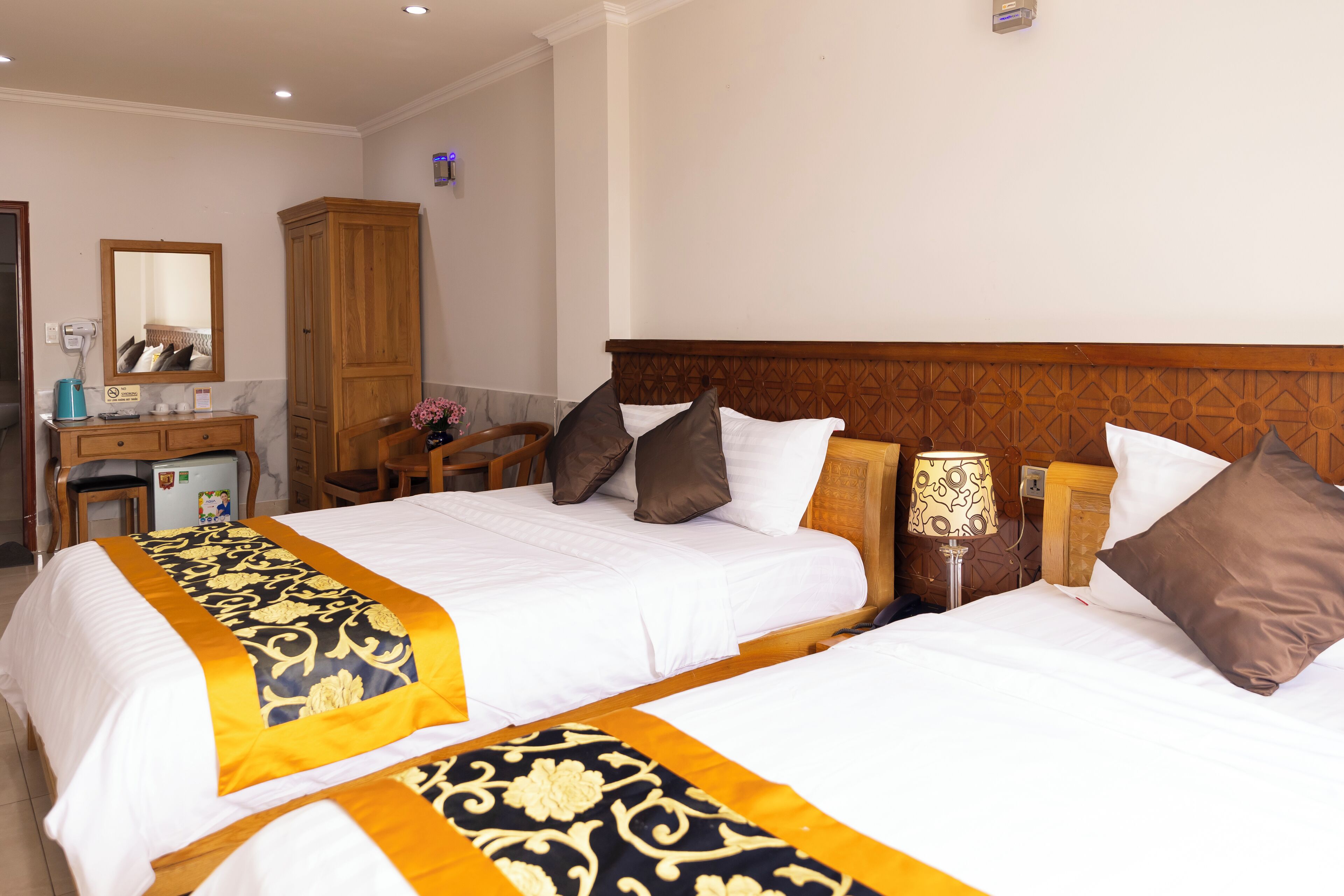 A hotel room with two beds, a wooden wardrobe, a desk with a mirror, and a small refrigerator.