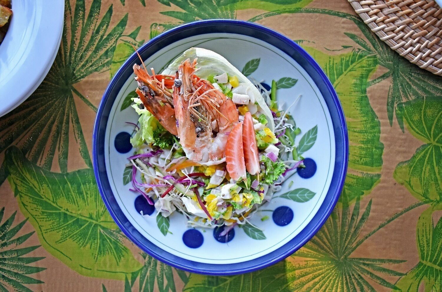 A bowl of salad with shrimp and vegetables on a tablecloth with a leaf pattern.