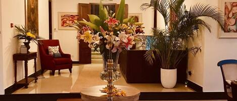 A lobby with a central table, a red armchair, a large potted plant, and a chandelier.
