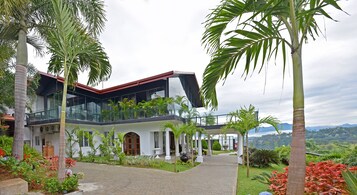 A modern two-story building with a glass balcony, surrounded by lush greenery and palm trees.