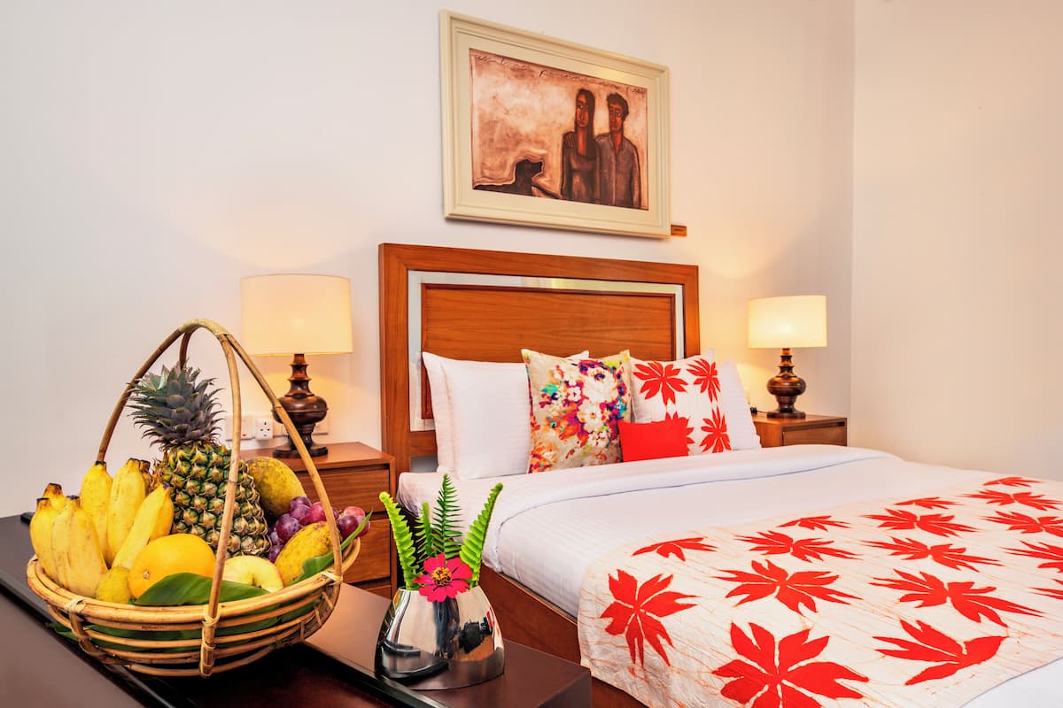 Ein Hotelzimmer mit einem hölzernen Kopfteil, einem Bett mit einer rot-weißen blumigen Tagesdecke, zwei Nachttischlampen, einem Nachttisch mit Obstkorb und Blumenvase sowie einem Gemälde an der Wand.