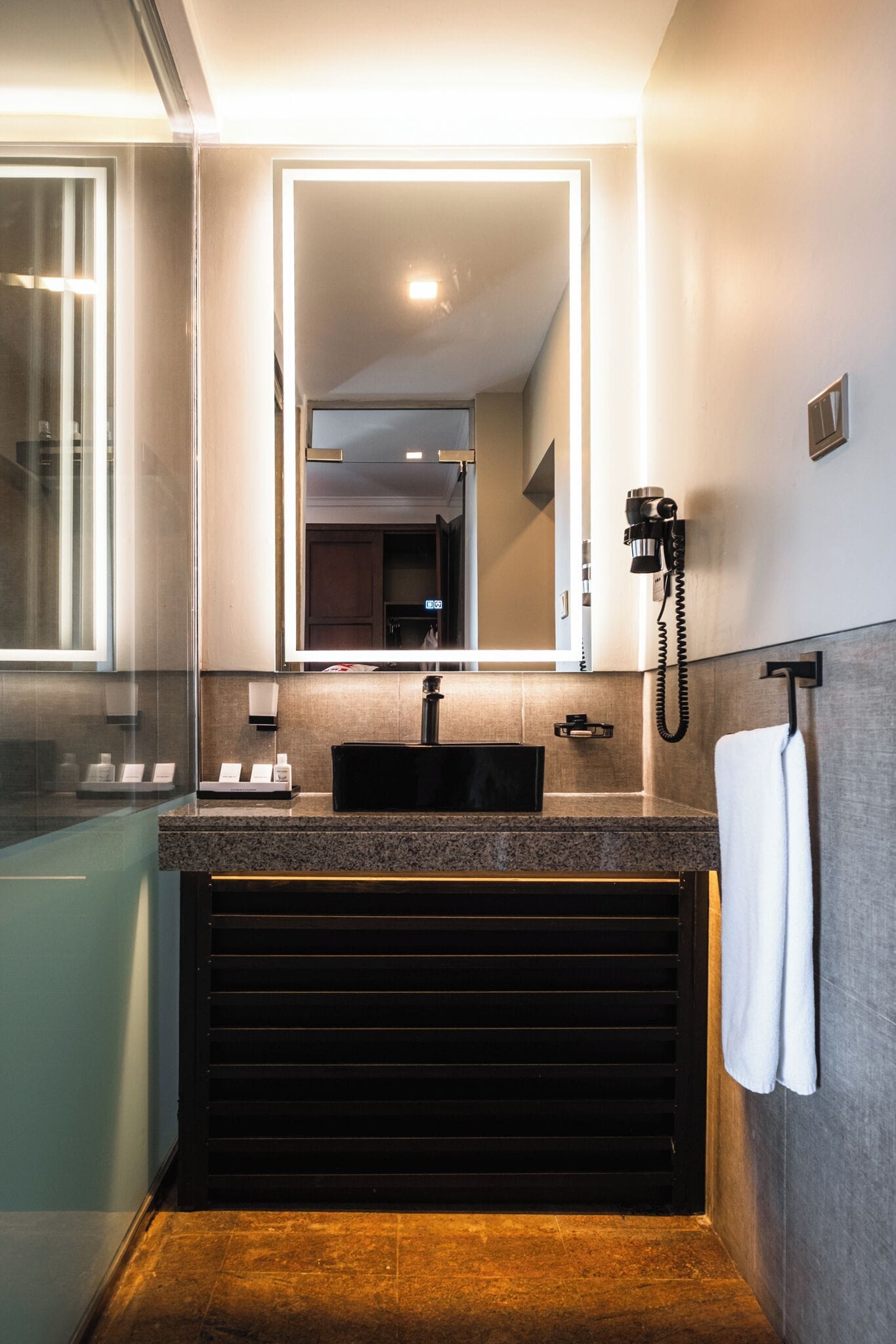 Un baño moderno con encimera de granito, lavamanos negro, espejo con iluminación LED, teléfono y toallero.