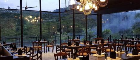 Un restaurante con mesas dispuestas para comer, amplios ventanales que ofrecen vistas al exterior y lámparas colgantes.