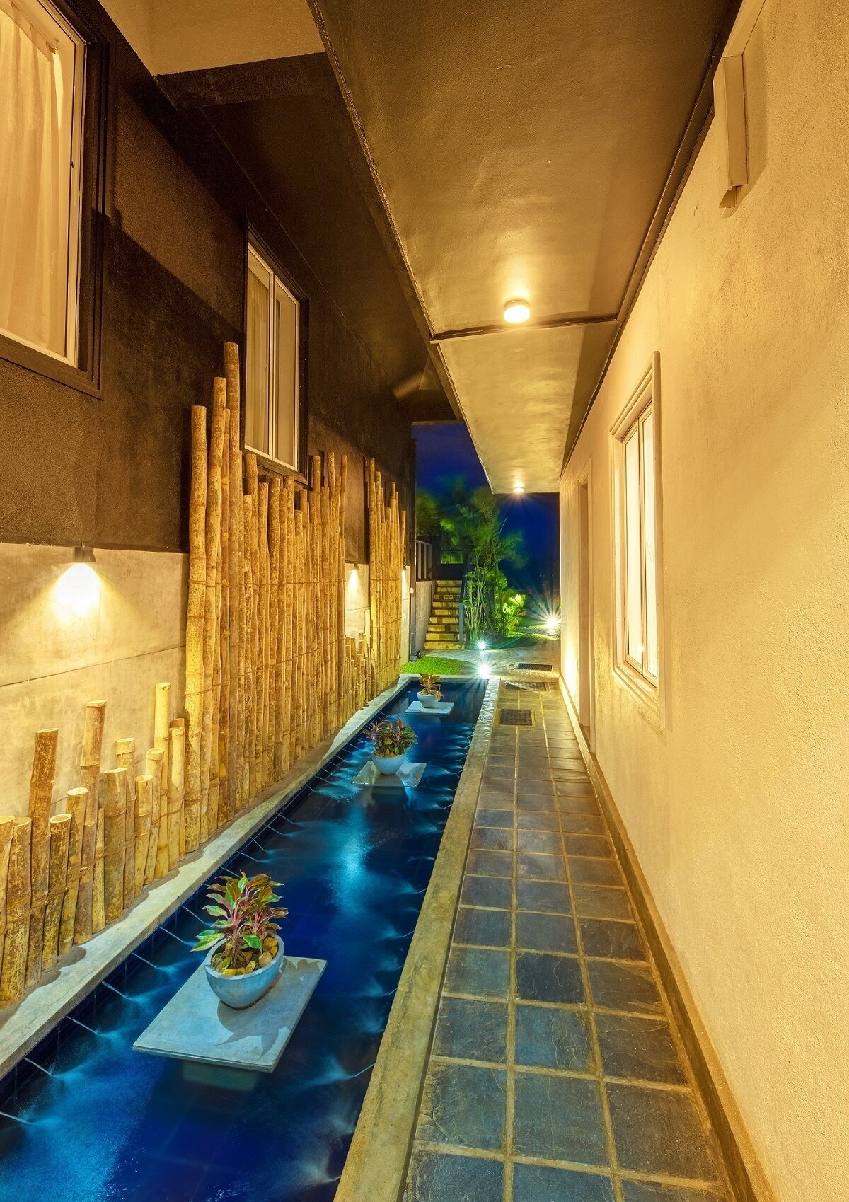 A narrow corridor with a reflective pool, bamboo wall, and potted plants.