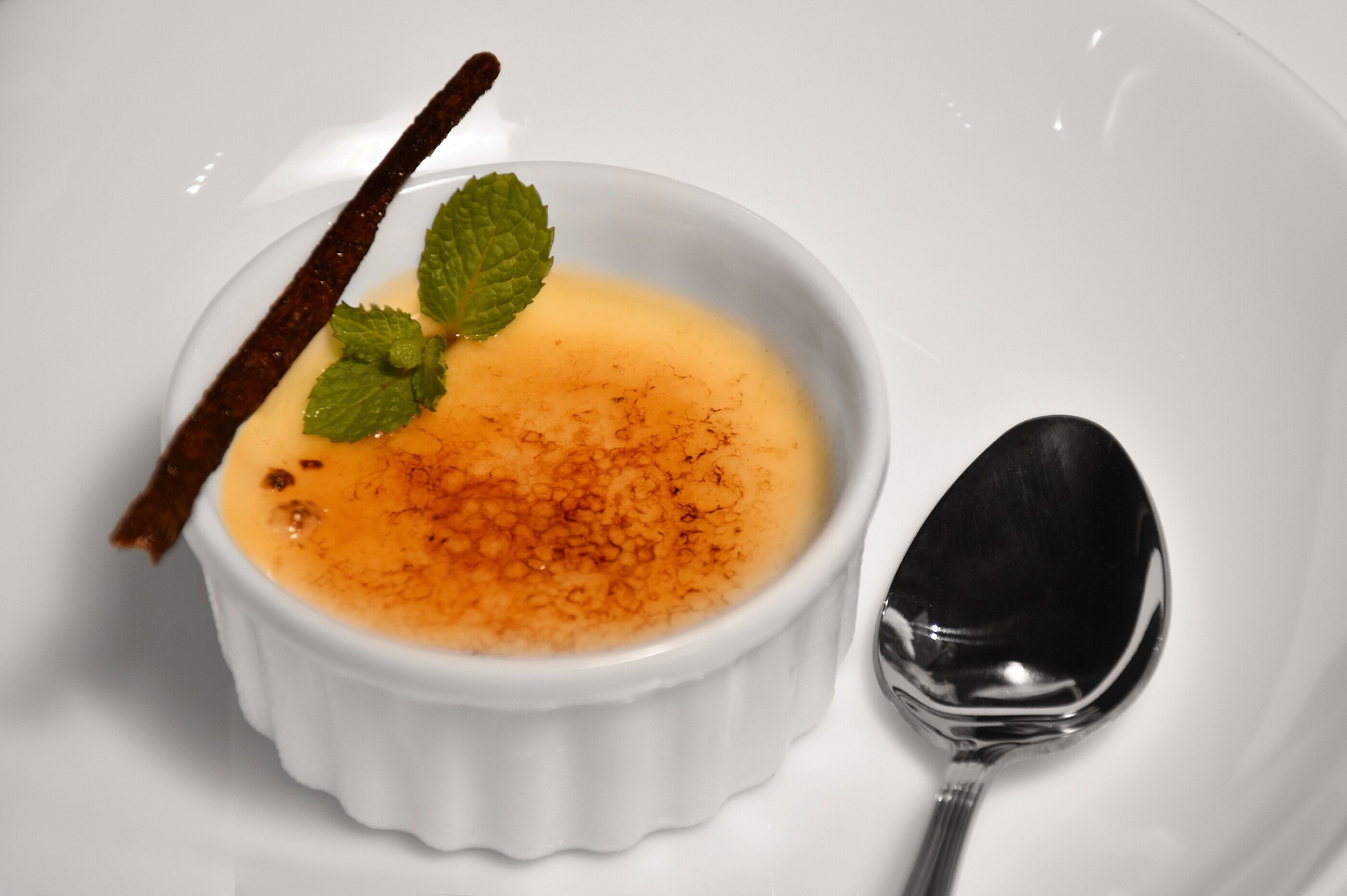 A white bowl of crème brûlée with a caramelized top, garnished with a mint leaf and a cinnamon stick, accompanied by a spoon.