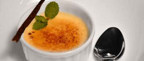 A white bowl of crème brûlée with a caramelized top, garnished with a mint leaf and a cinnamon stick, accompanied by a spoon.
