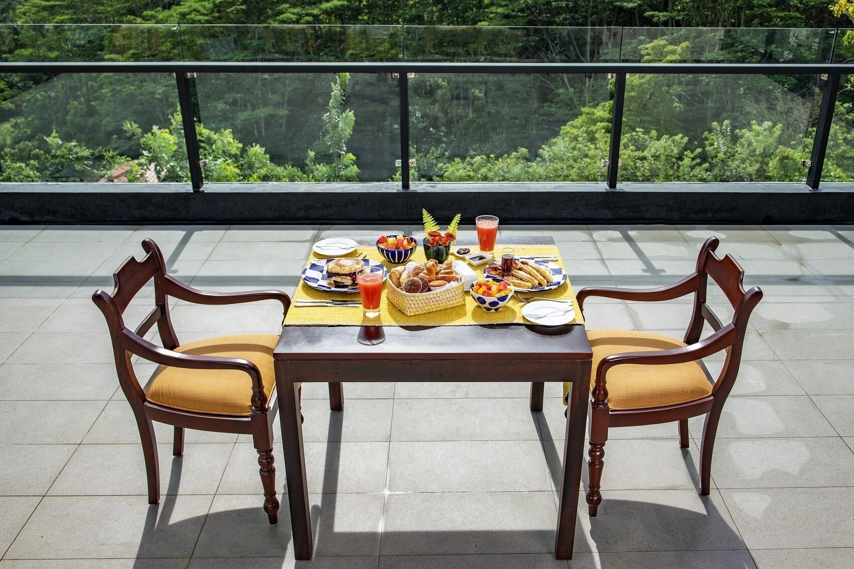Una mesa de comedor con comida servida, dos sillas y una barandilla de vidrio con vista a un área boscosa.