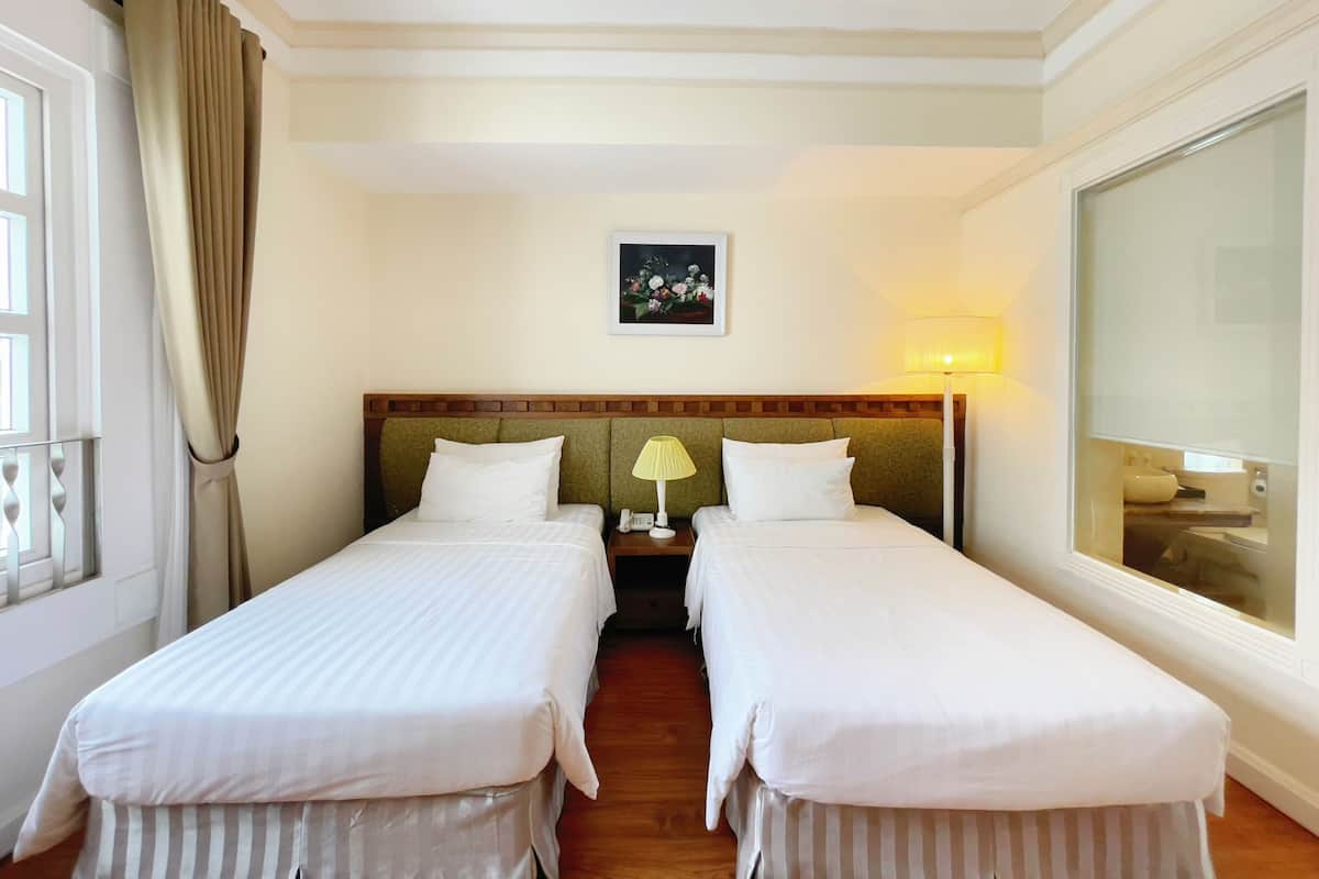 Une chambre d’hôtel avec deux lits simples, une table de chevet avec une lampe, un tableau encadré au mur et une grande fenêtre.