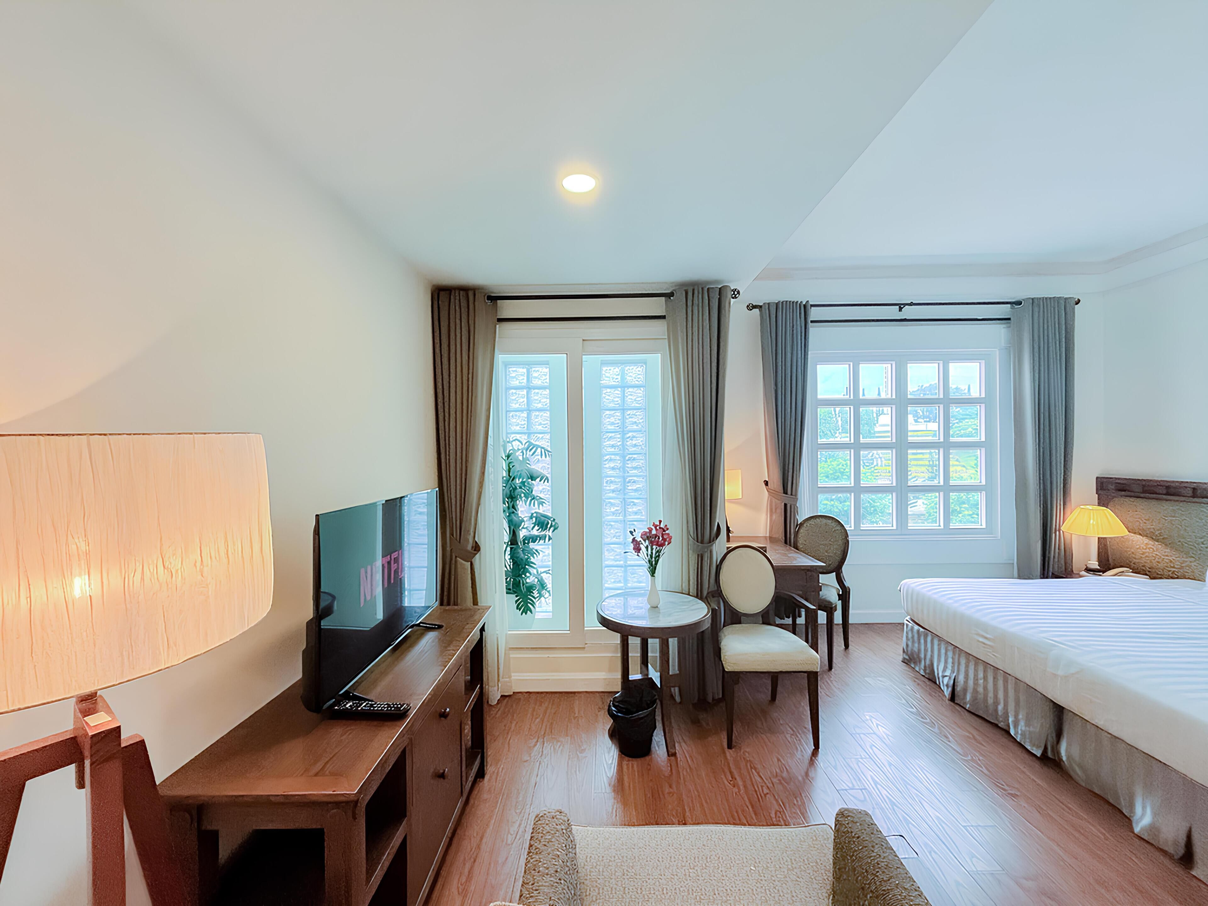 A hotel room with a bed, a desk with a TV, a chair, a small table with flowers, and a window with curtains.