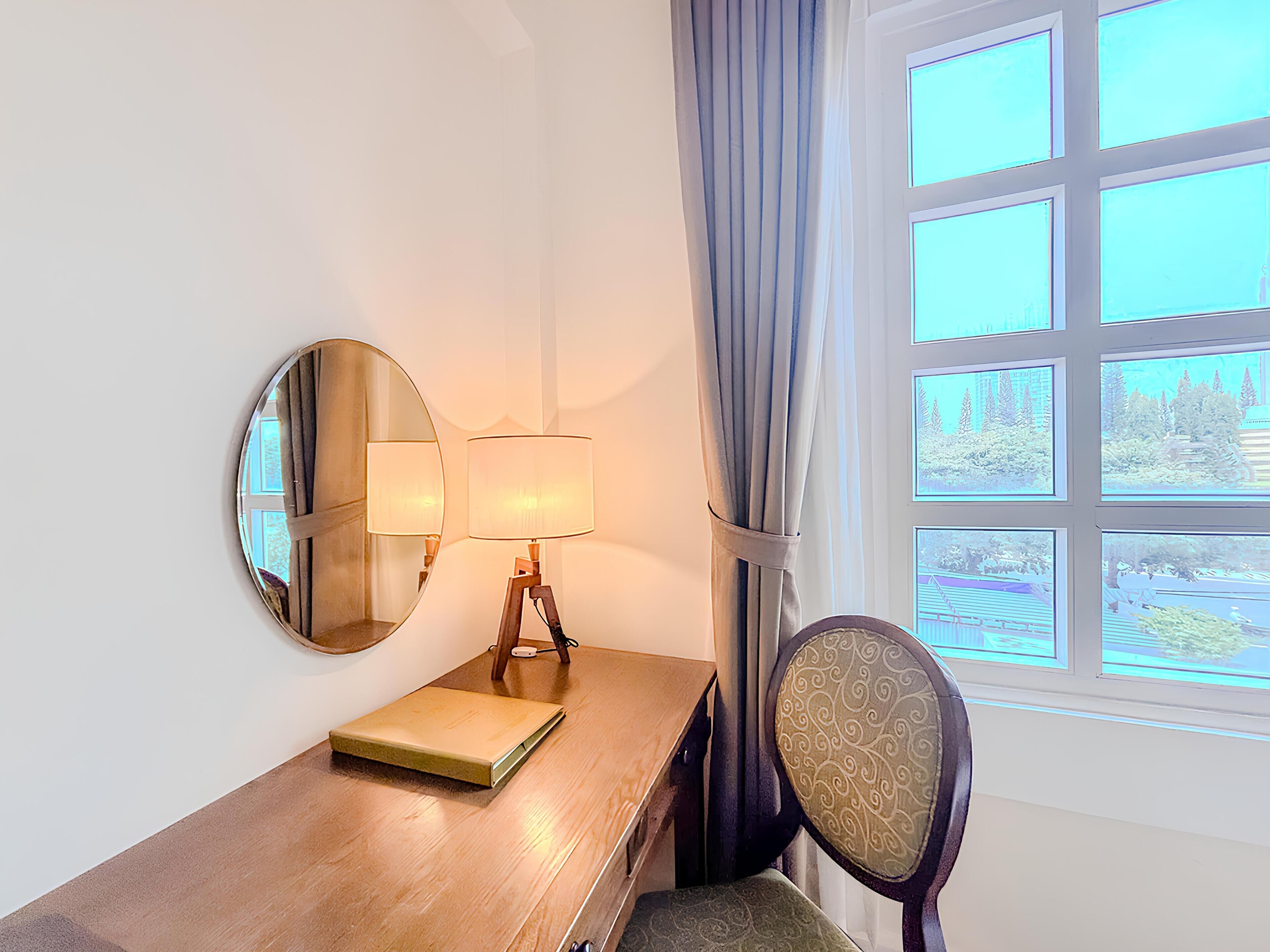 A room with a wooden desk, a round mirror, a lamp, a chair, and a window with a view of trees and a building.