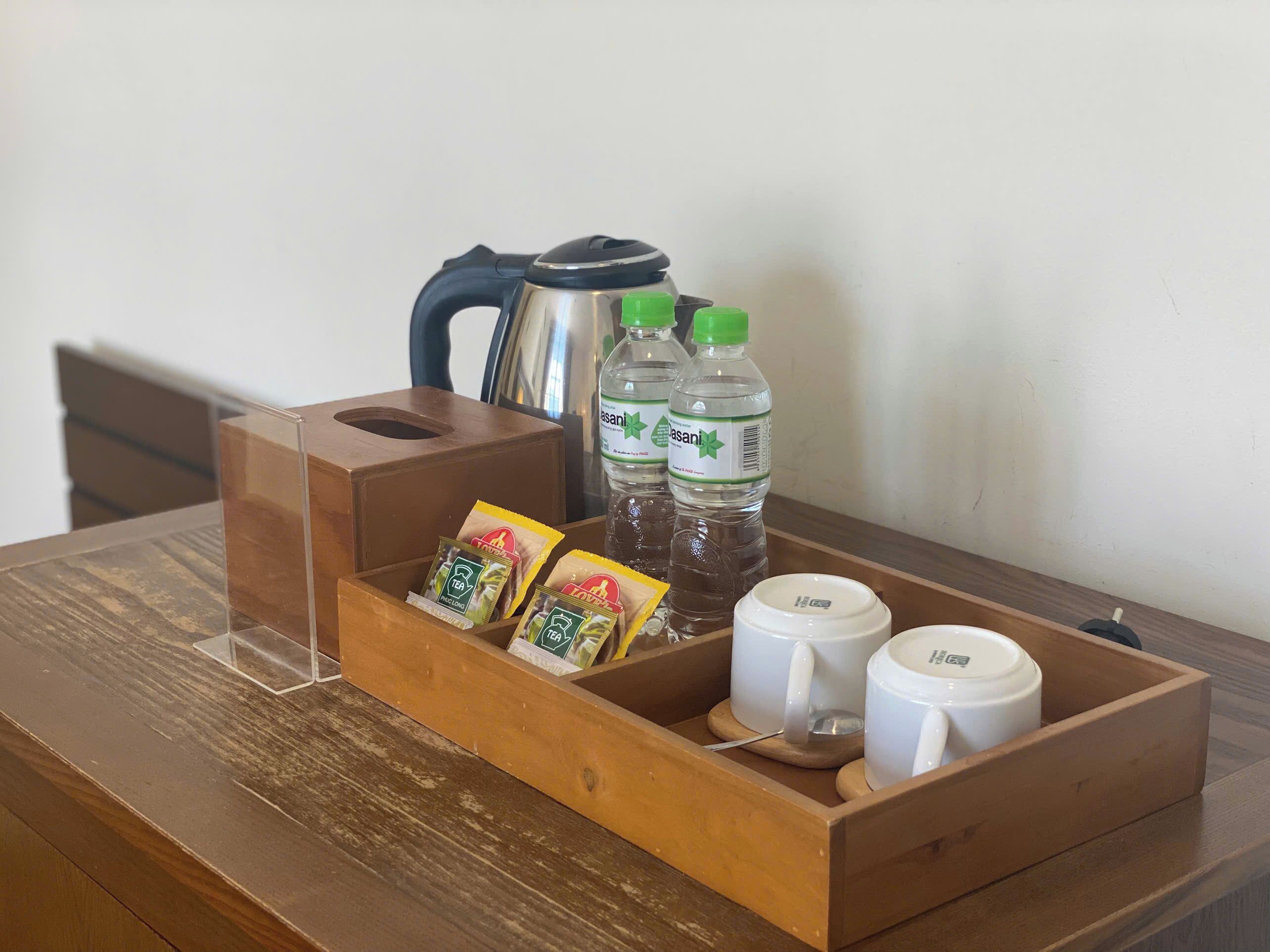 A wooden tray with tea packets, two water bottles, a kettle, and two white cups on a wooden table.