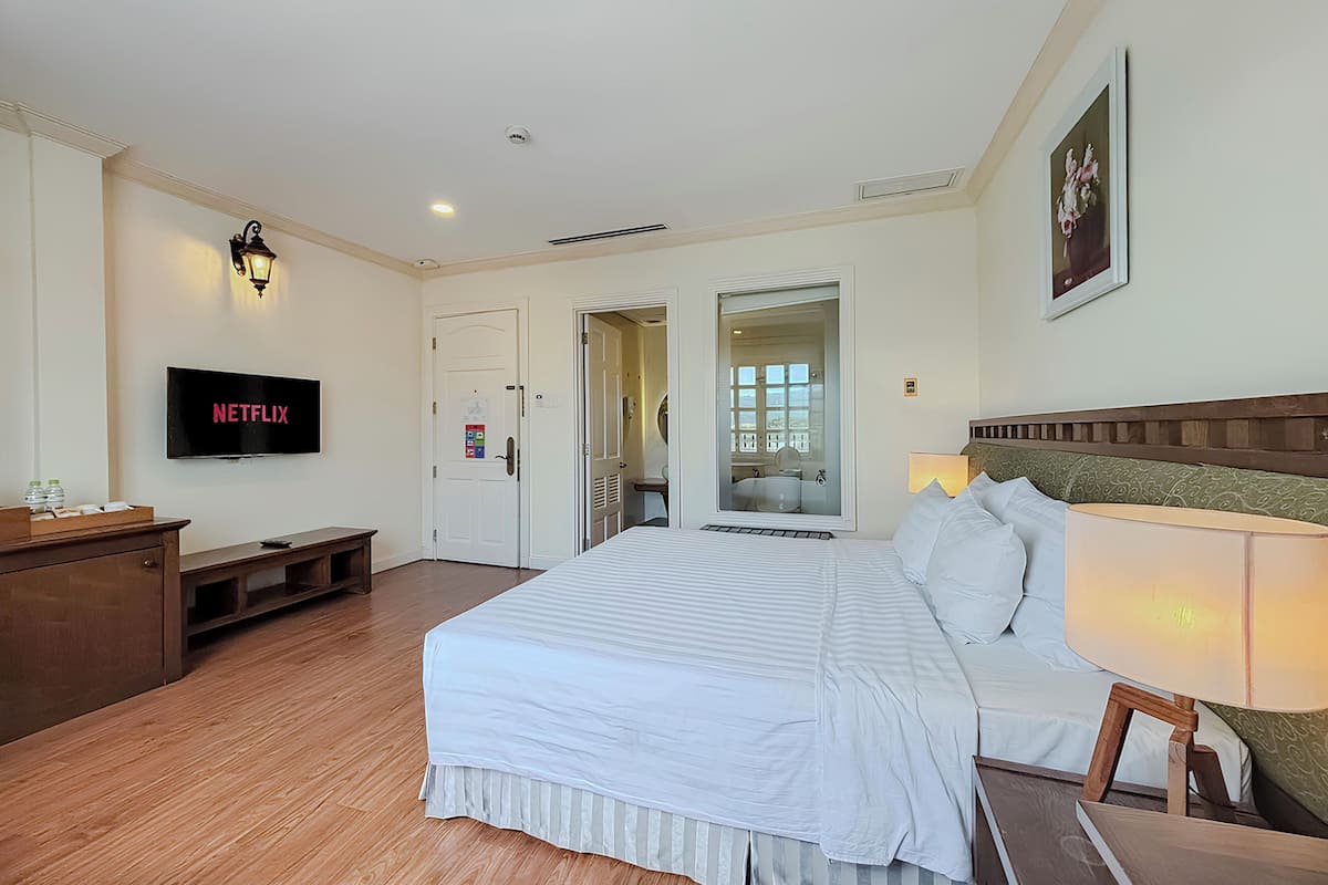 Une chambre d’hôtel avec un grand lit, un téléviseur fixé au mur, un banc en bois et une porte donnant sur une salle de bain.