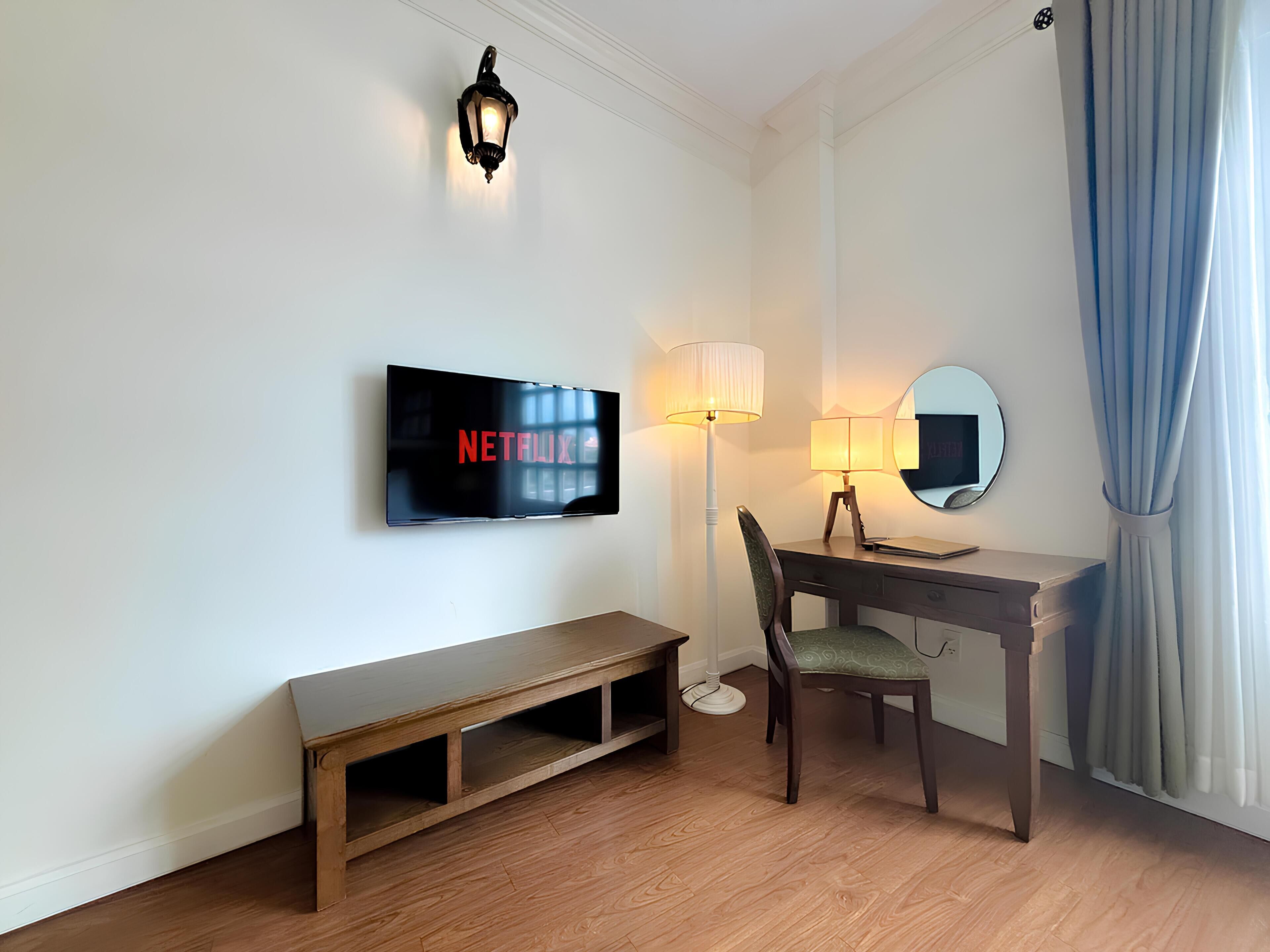 A hotel room with a flat-screen TV displaying 'NETFLIX', a desk with a lamp, a chair, a bench, a mirror, and a window with curtains.