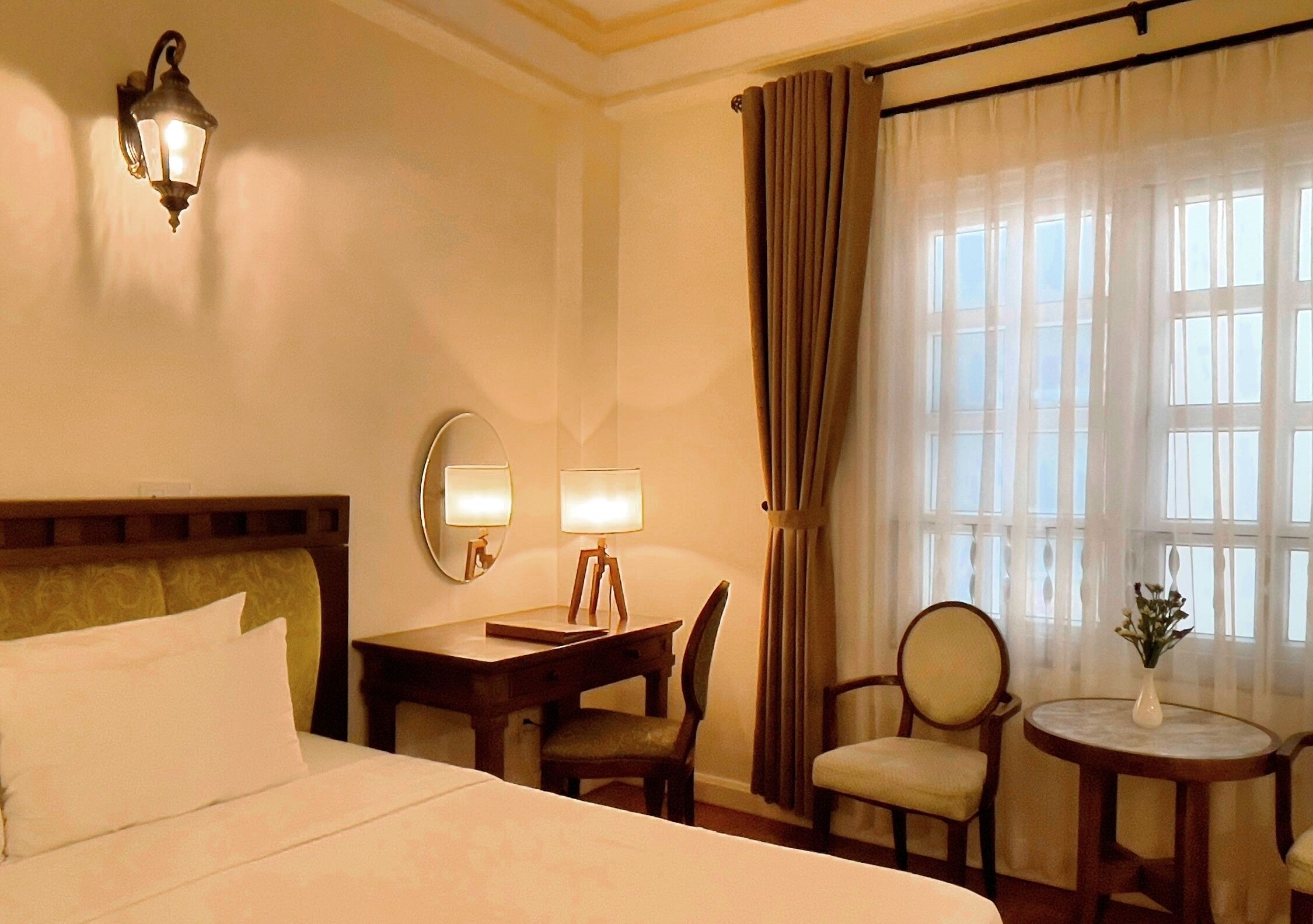 Ein Hotelzimmer mit einem Bett, zwei Nachttischlampen, einem Schreibtisch mit Lampe, einem Sessel, einem runden Tisch mit einer Vase und einem Fenster mit Vorhängen.