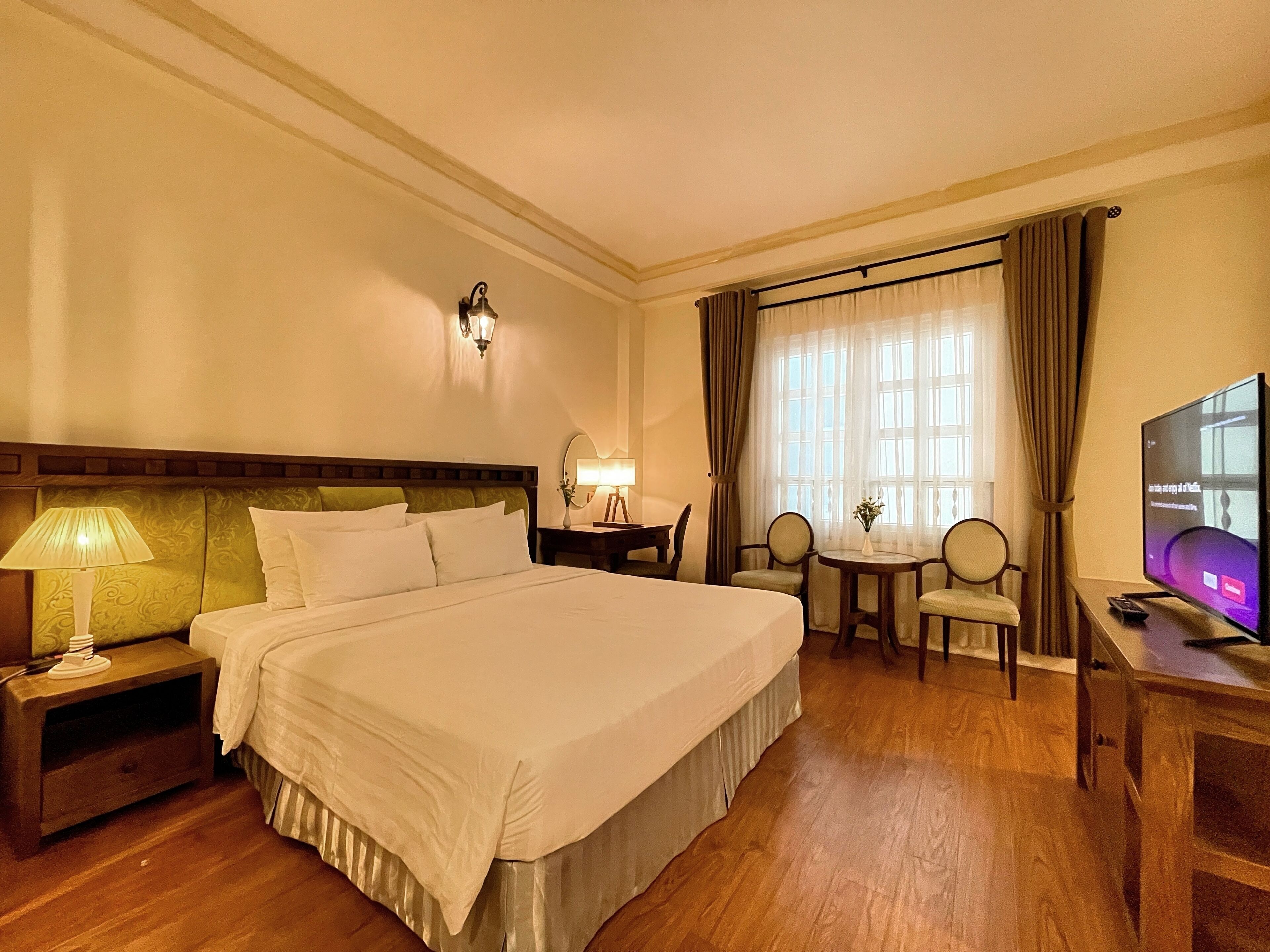 A hotel room with a large bed, a desk with a television, a small table with chairs, and a window with curtains.