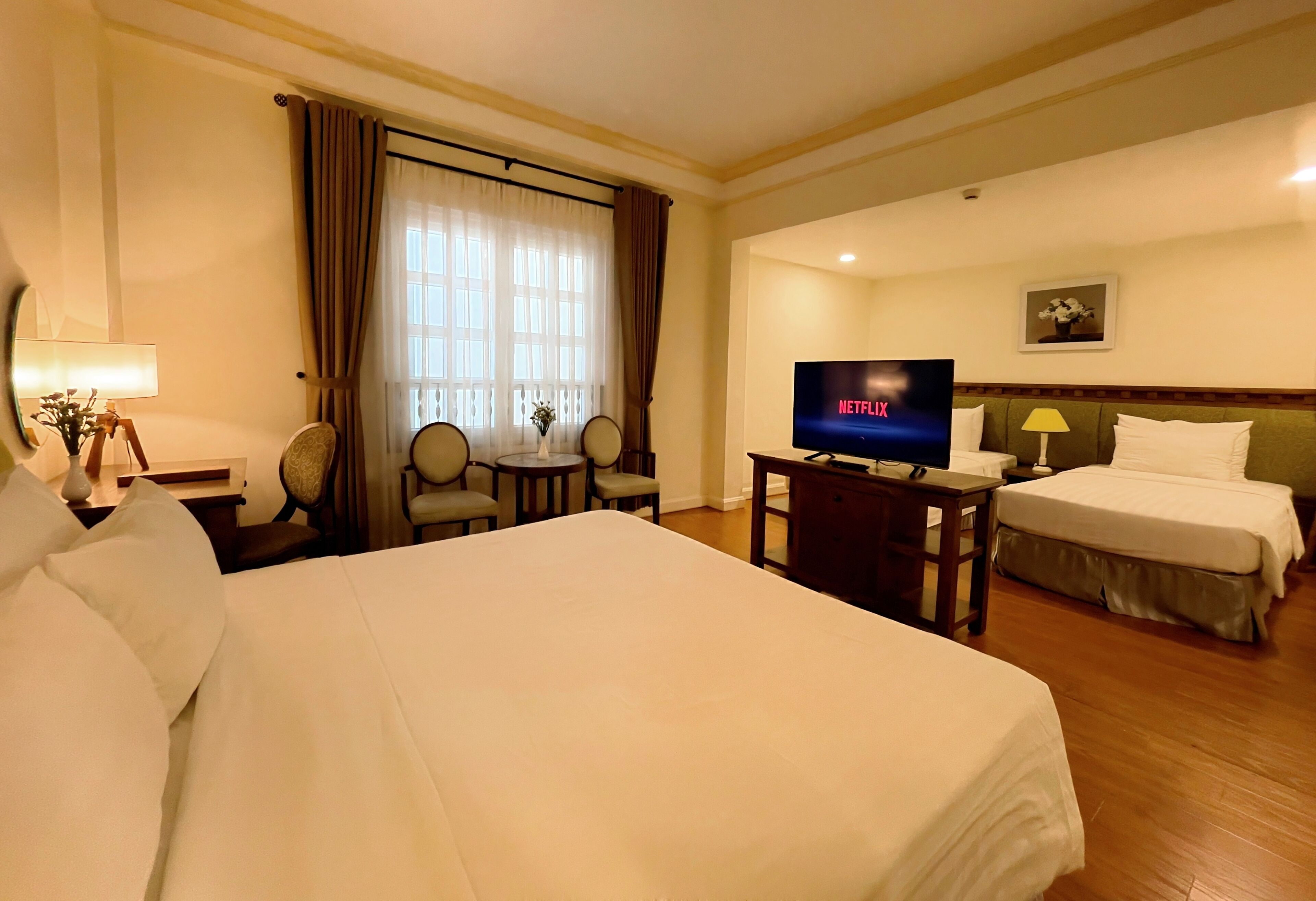 Hotelkamer met groot bed, televisie, bureau met lamp, twee stoelen, klein tafeltje en raam met gordijnen.