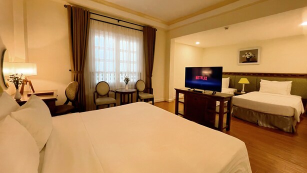 Ein Hotelzimmer mit einem großen Bett, einem Fernseher, einem Schreibtisch mit Lampe, zwei Stühlen, einem kleinen Tisch und einem Fenster mit Vorhängen.