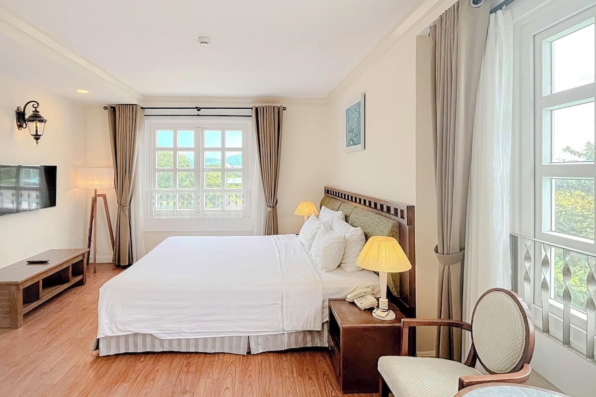 Une chambre d’hôtel avec un grand lit, des lampes de chevet, un banc en bois, une chaise et une vue sur la verdure par la fenêtre.