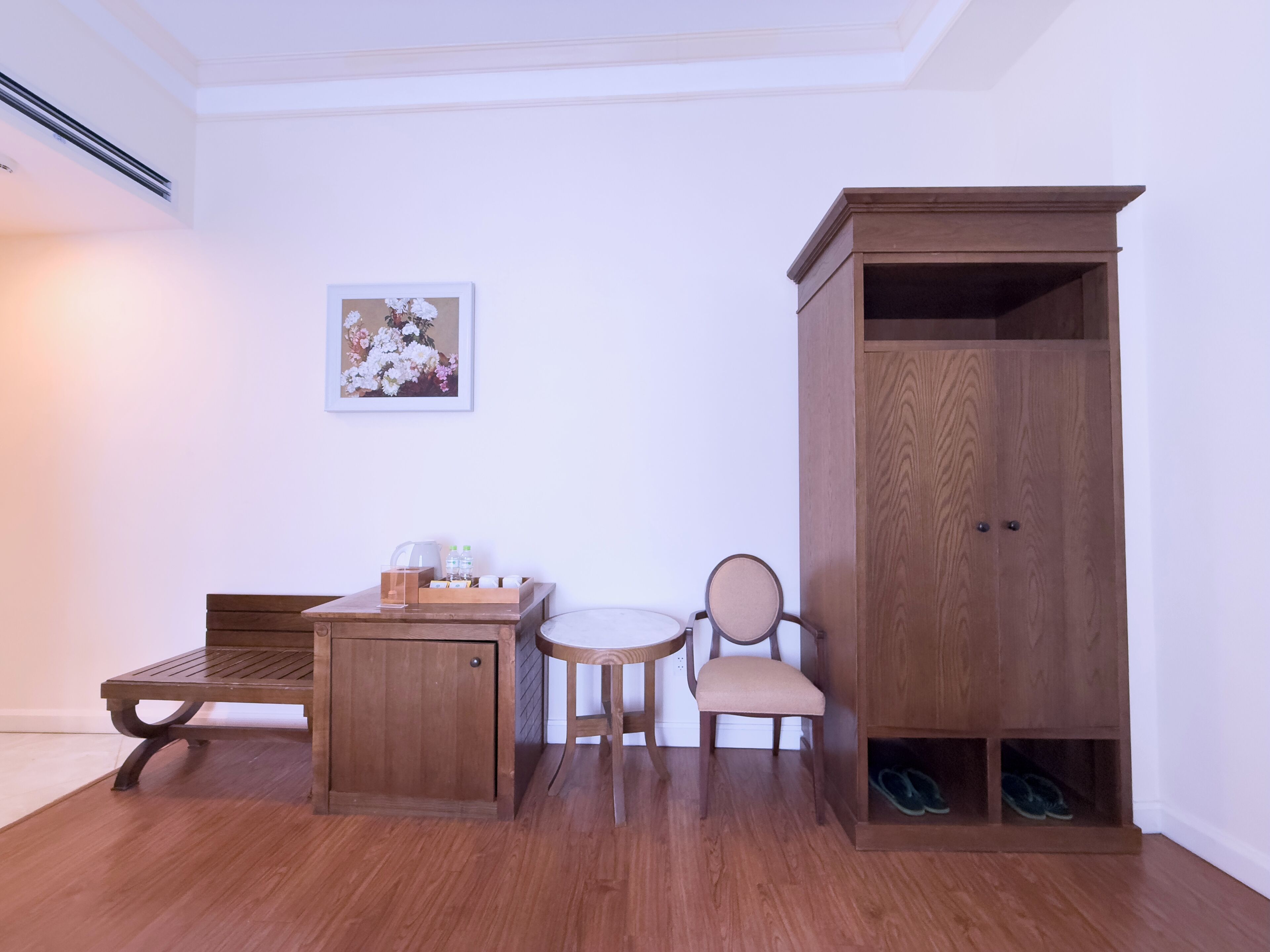 Una habitación con un banco de madera, una mesita con una tetera y botellas, una silla, un armario de madera y un par de zapatos en una caja de zapatos.