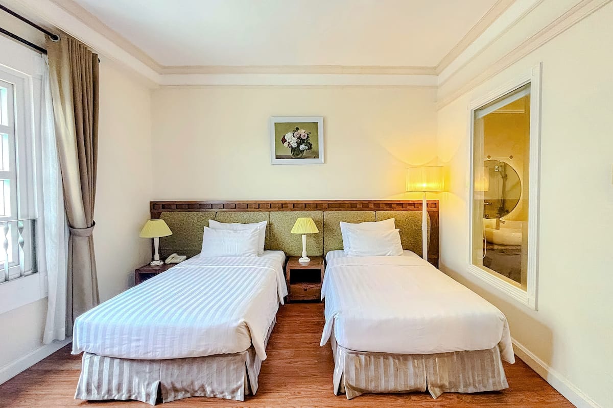 Une chambre d’hôtel avec deux lits simples, une tête de lit en bois, un tableau au mur, une fenêtre avec des rideaux et une salle de bain visible à travers une porte en verre.