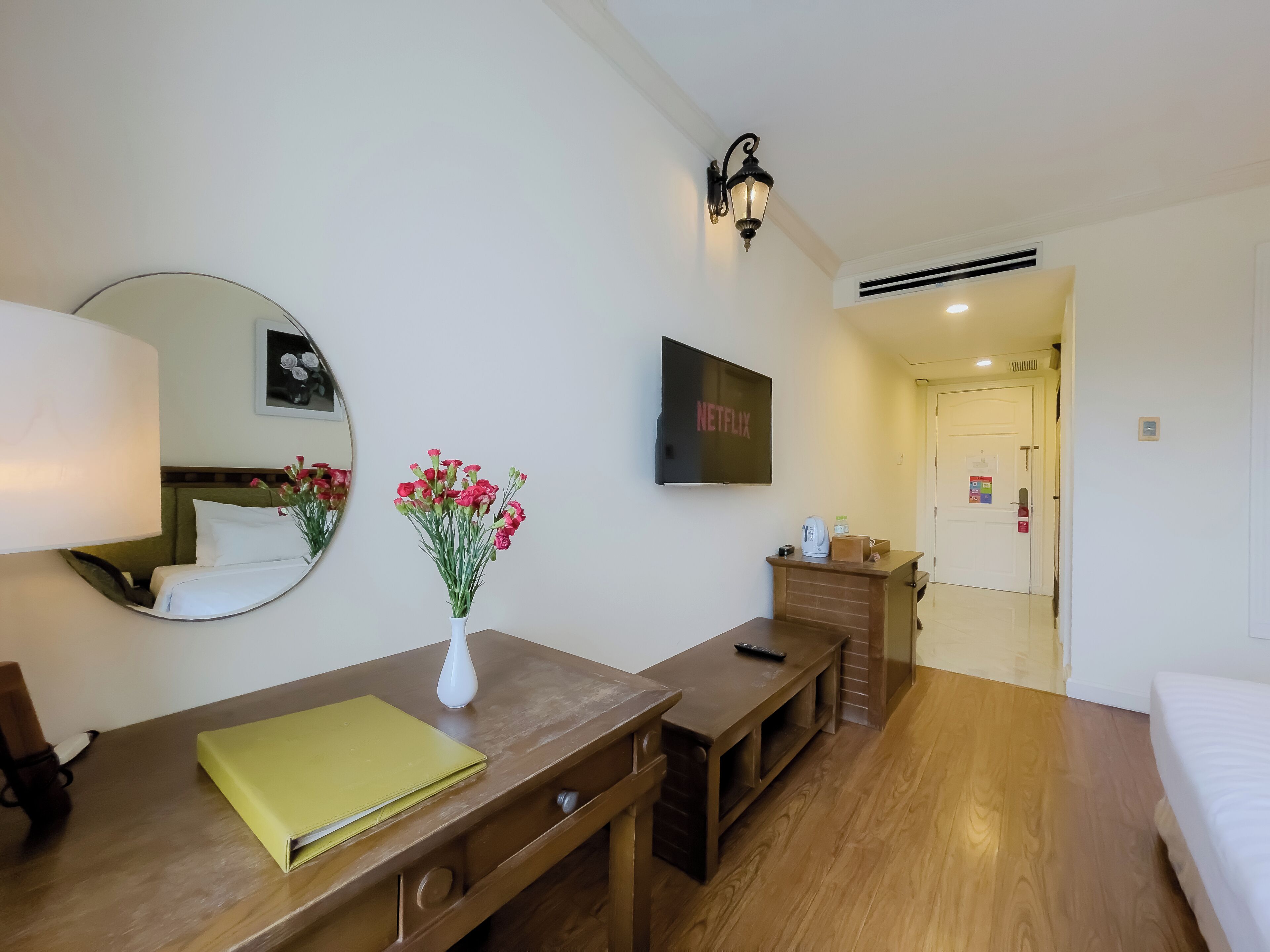A hotel room with a bed, a desk with a laptop, a mirror, a vase with flowers, a television, and a door leading to another room.