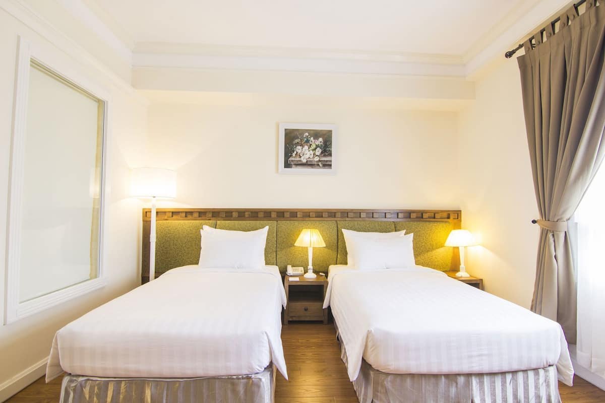 Une chambre d’hôtel avec deux lits simples, une tête de lit ornée d’un motif, des lampes de chevet, un tableau encadré au mur et un rideau à la fenêtre.