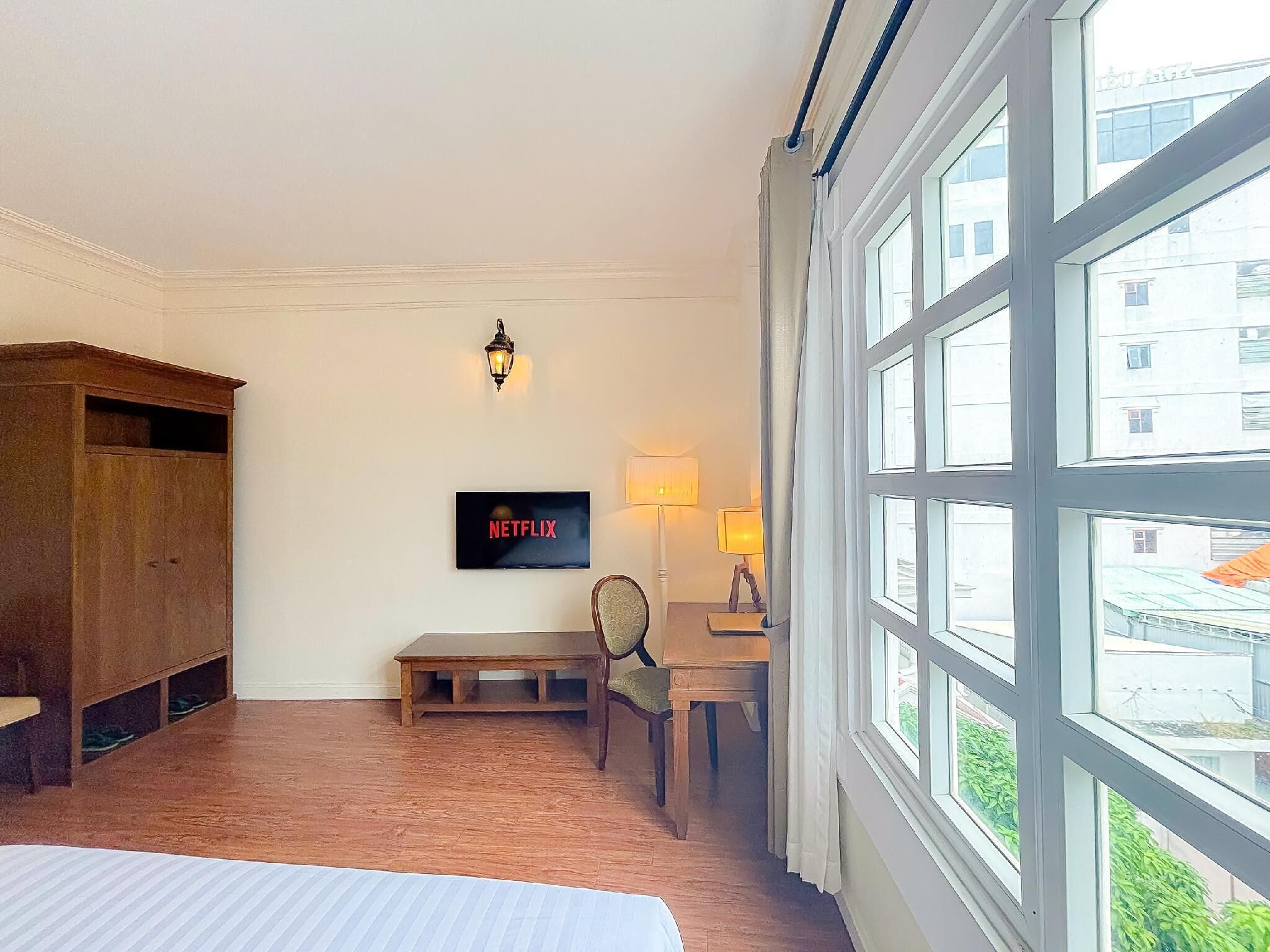A hotel room with a large window, a wooden wardrobe, a desk with a chair, a wall-mounted TV, and a lamp.