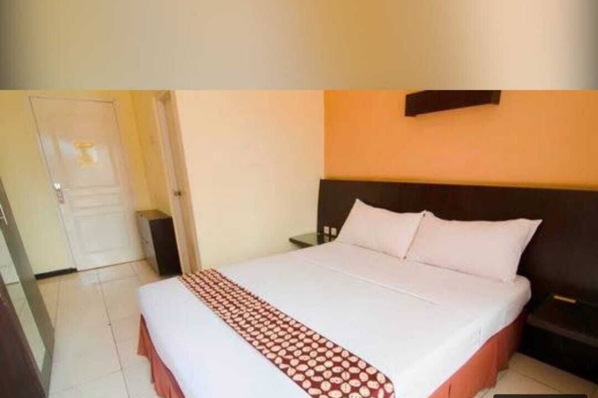 A hotel room with a bed, a nightstand, a wall-mounted shelf, and a door.