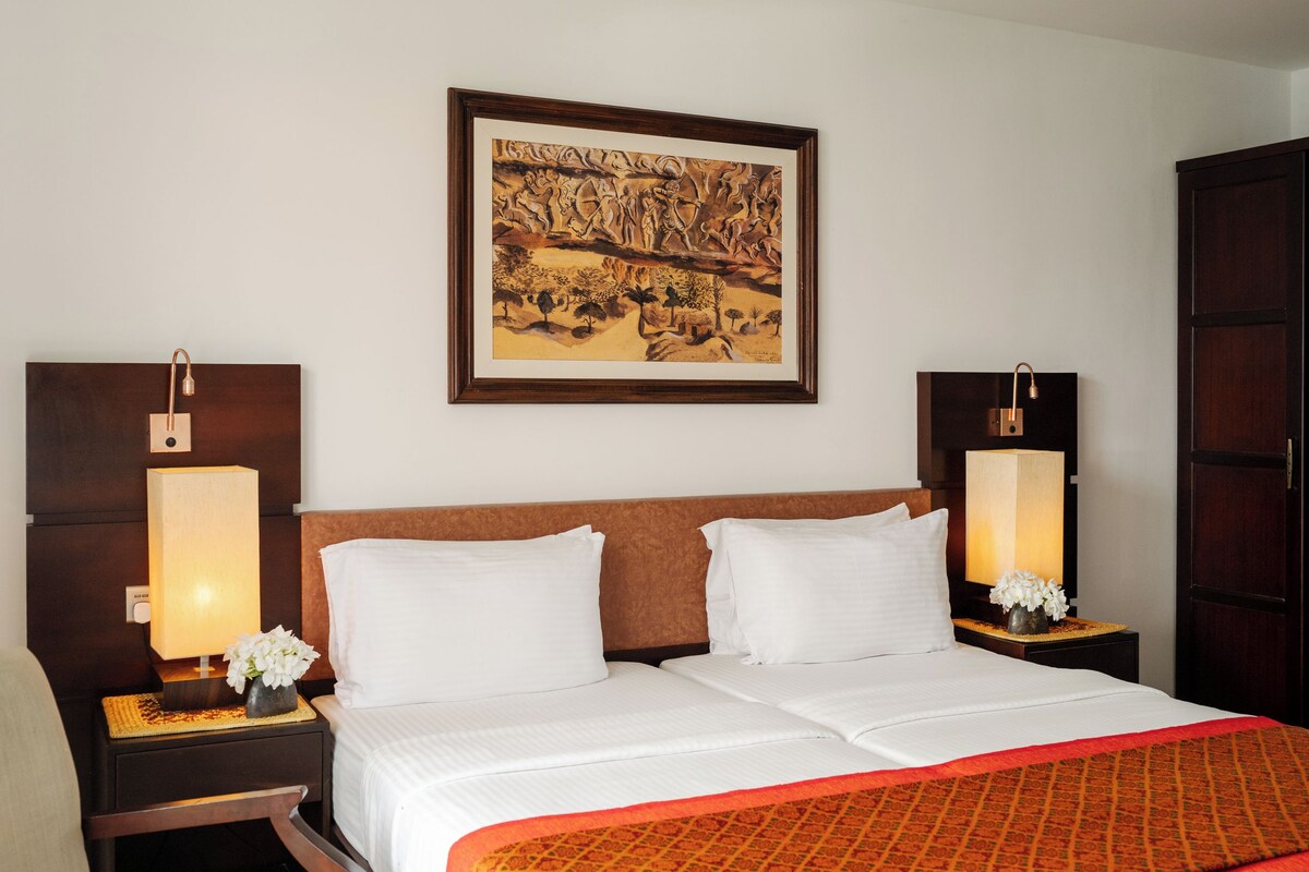 Una habitación de hotel con una cama grande, dos mesitas de noche con lámparas, una obra de arte enmarcada sobre la cama y un armario de madera.