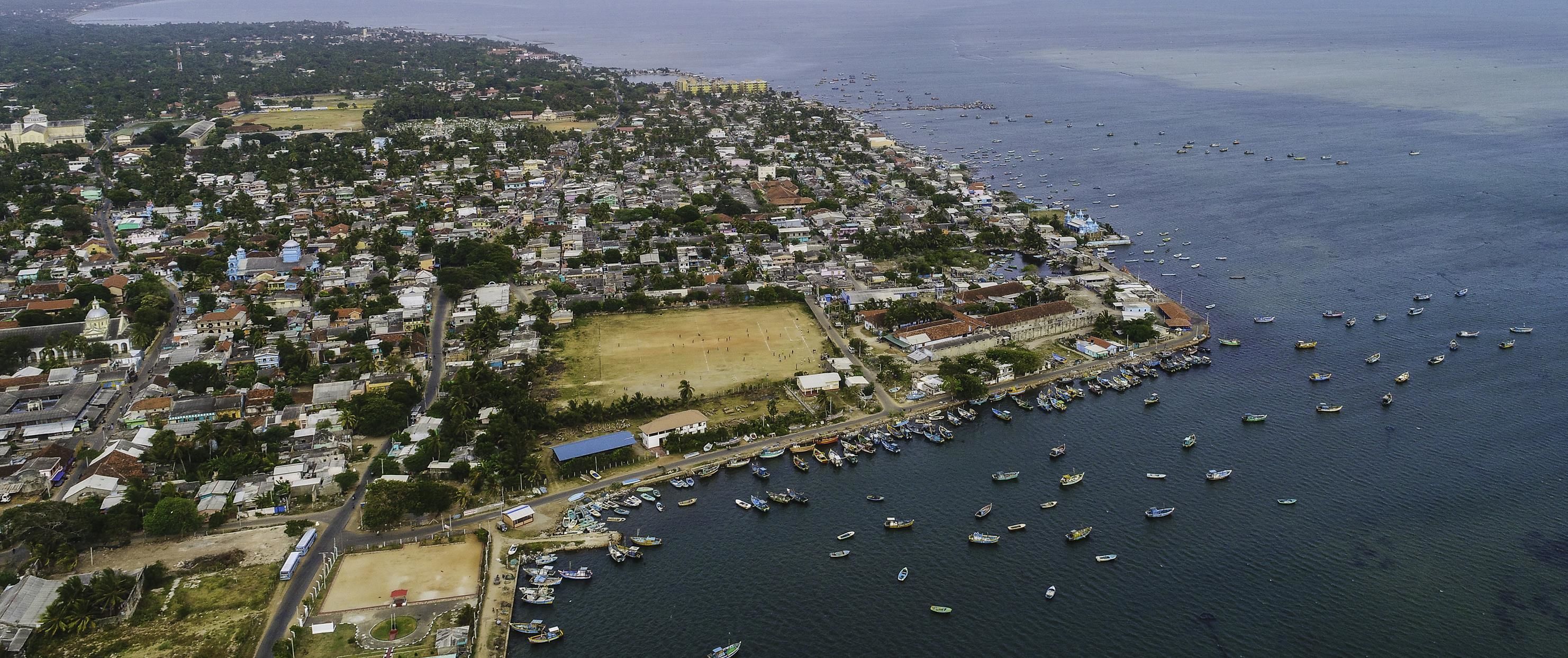 An aerial view of a coastal town with a marina filled with boats, surrounded by residential buildings and a large open field.