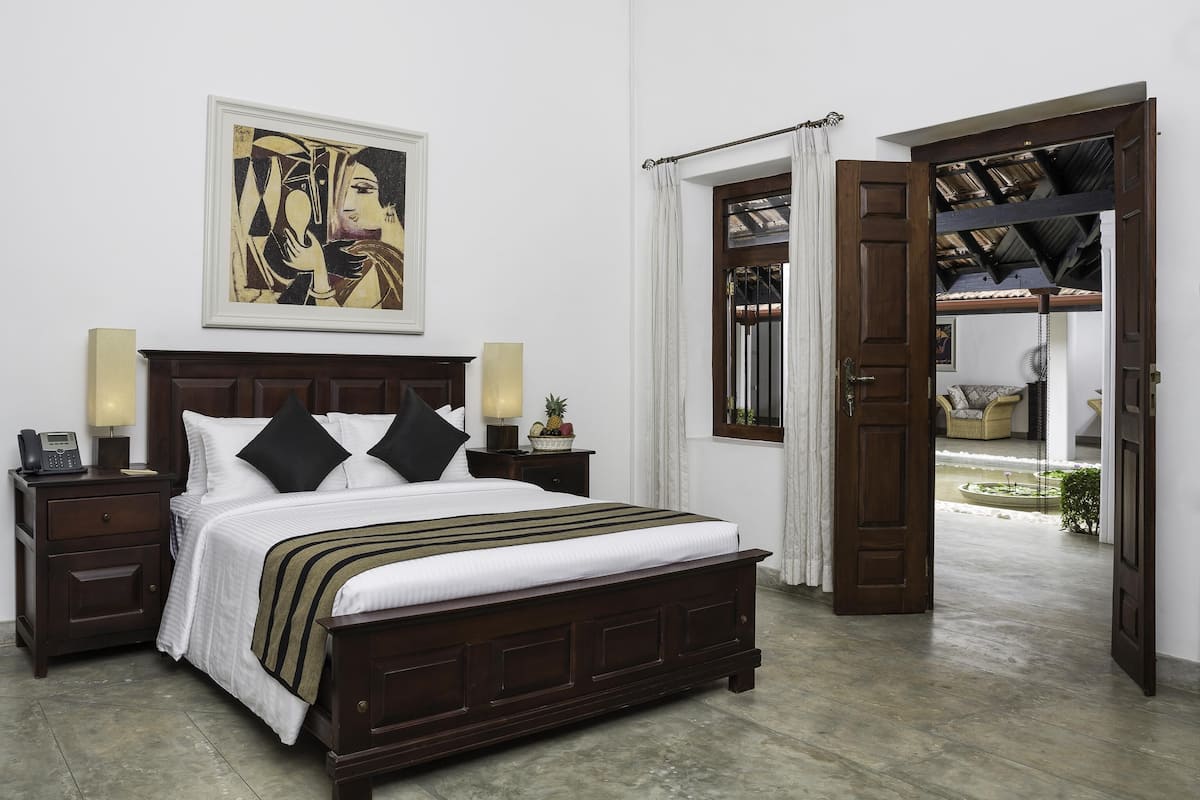 A bedroom with a wooden bed, a nightstand with a phone and lamp, a painting above the bed, and an open door leading to a balcony with a view of a garden.