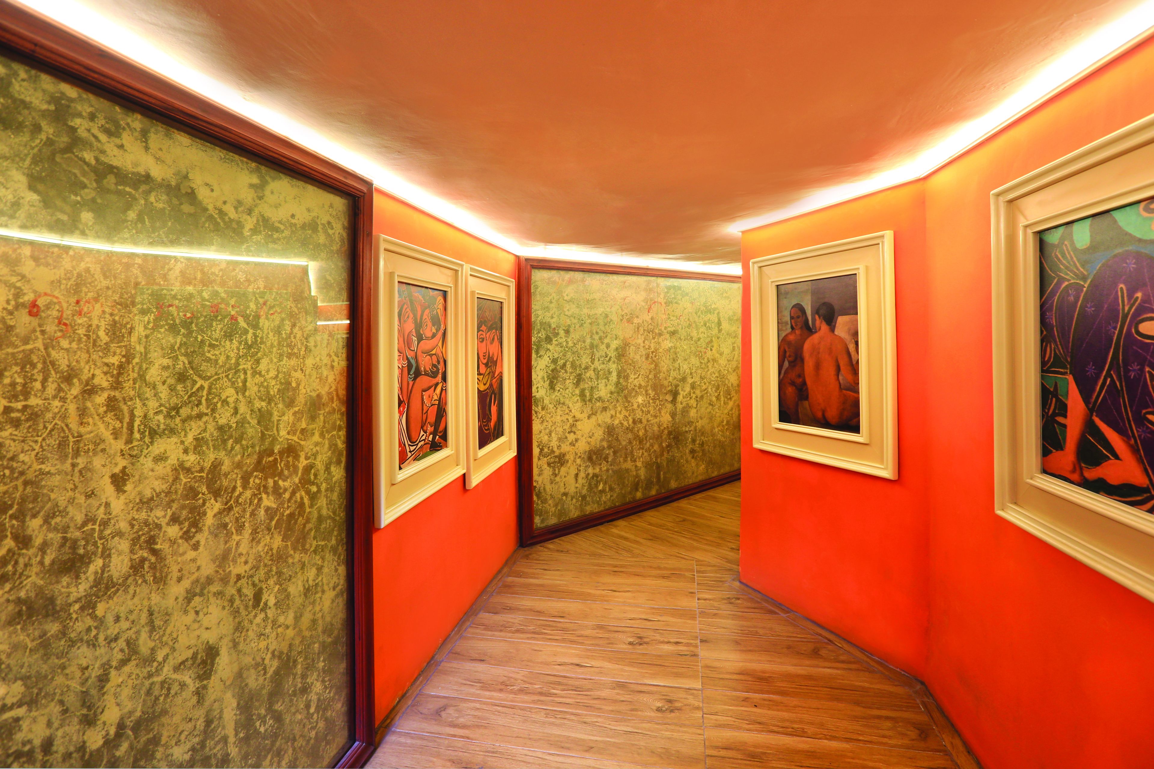 A hallway with orange walls and wooden flooring, featuring framed artworks on the walls.