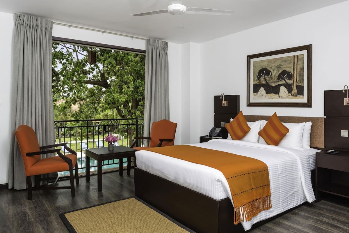 A hotel room with a bed, two chairs, a desk, a phone, a painting, and a view of greenery.