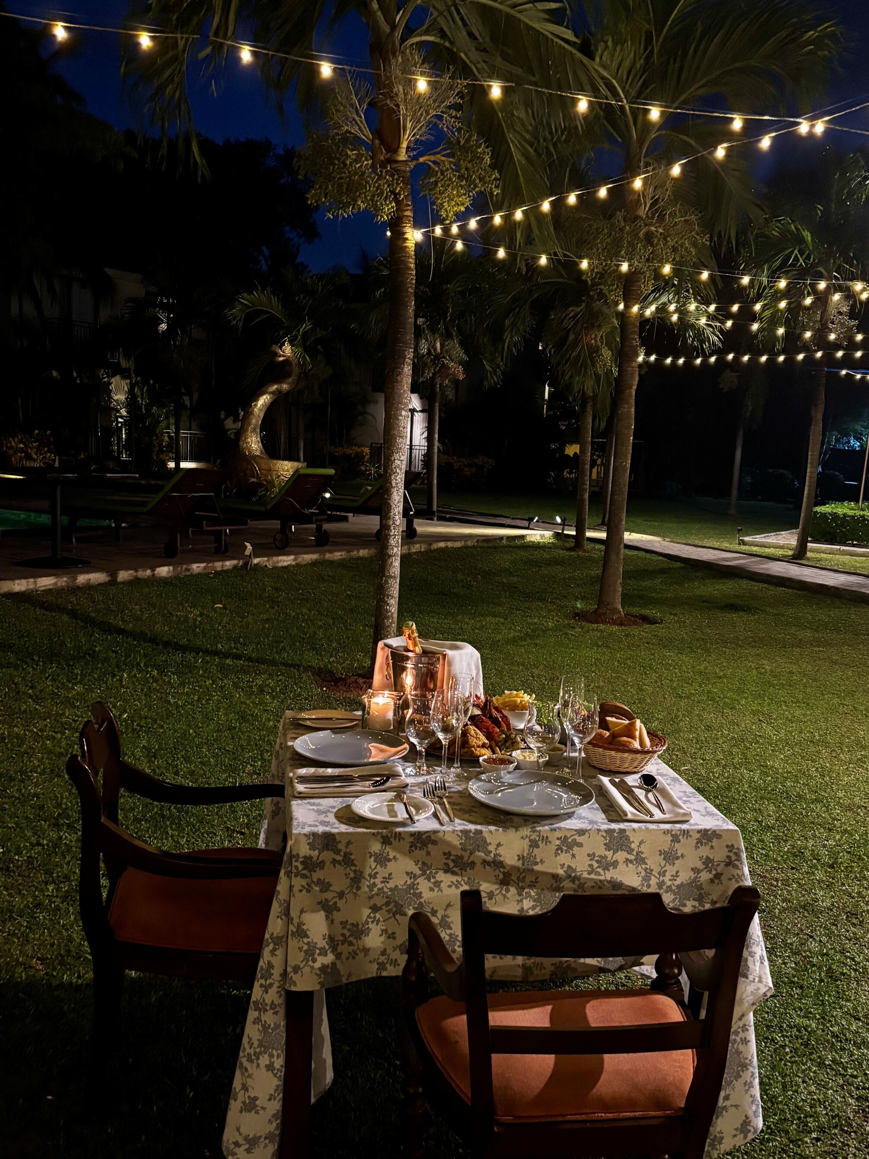 Mobiliario de comedor al aire libre con una mesa dispuesta para dos personas, rodeado de palmeras y luces colgantes.