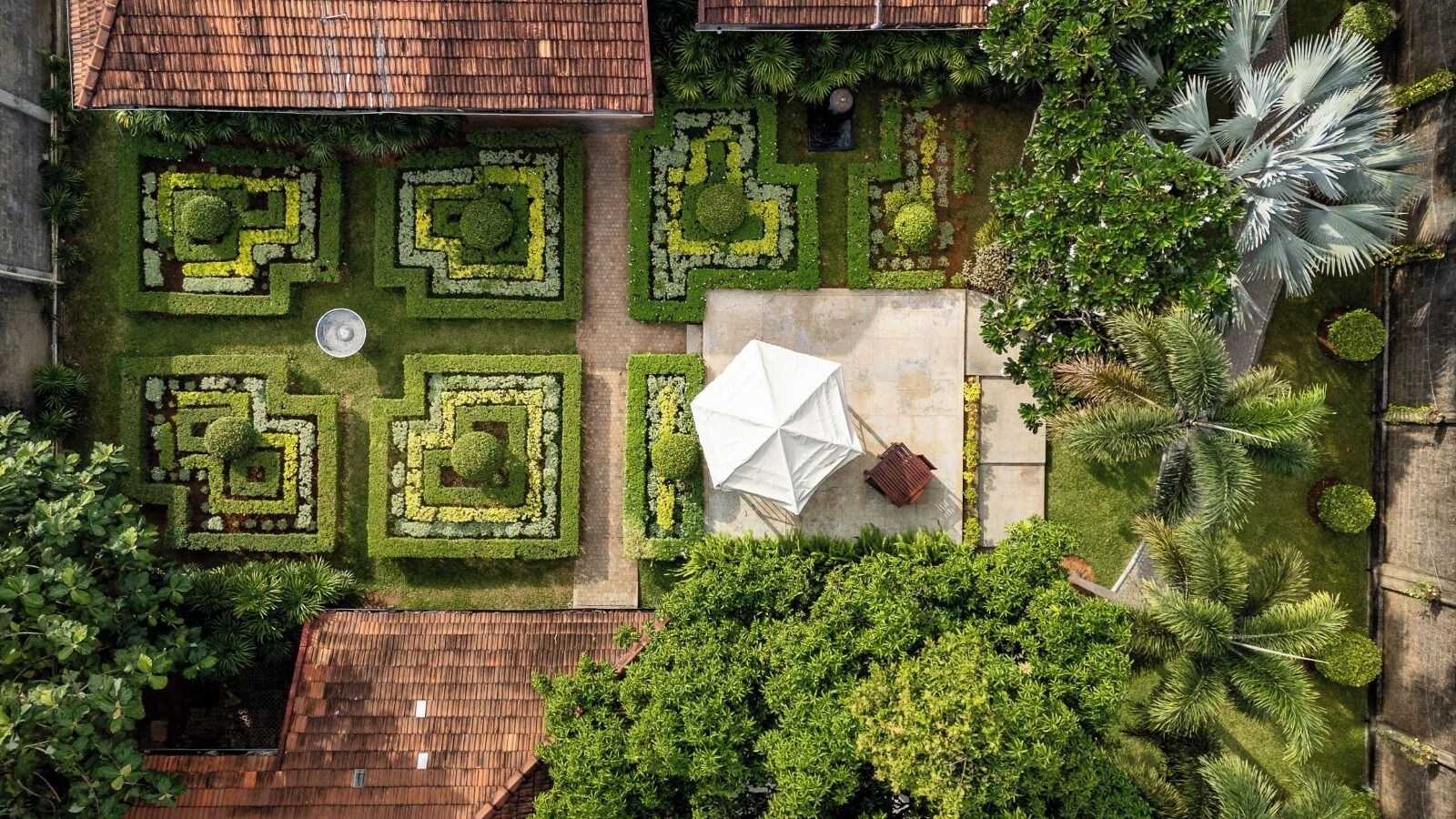 Vista aérea de um pátio com jardins de design geométrico, um guarda-sol branco e árvores ao redor.