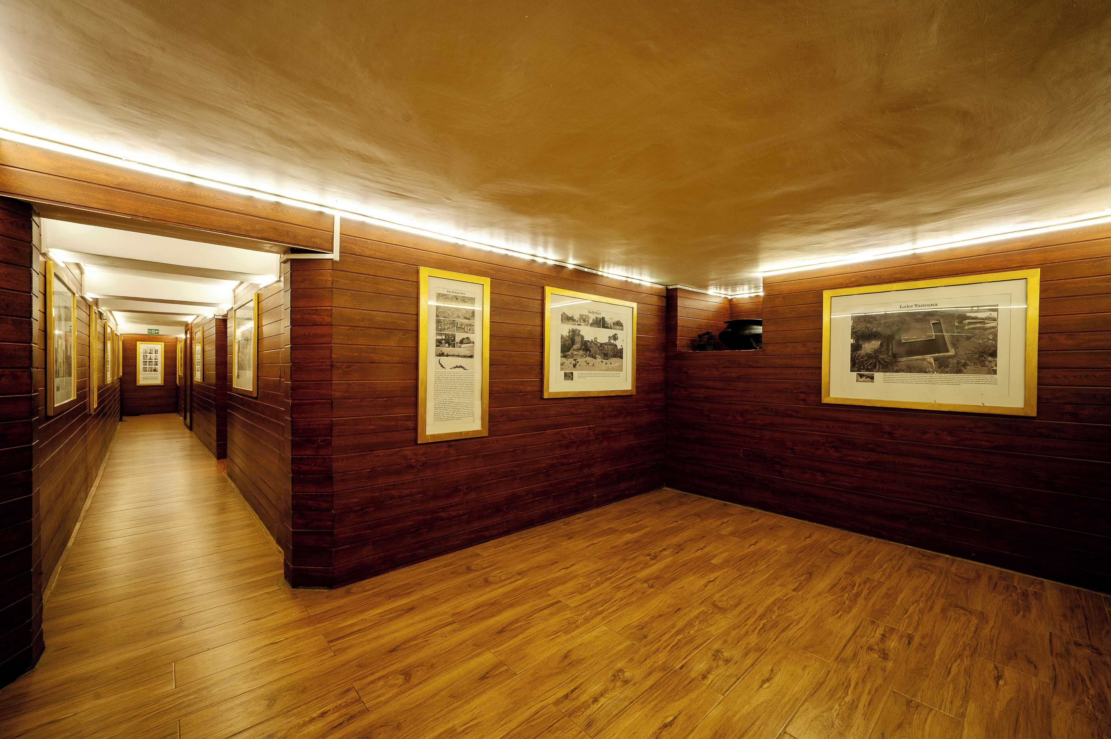 A hallway with wooden walls and floors, framed pictures on the walls, and a wooden door at the end.