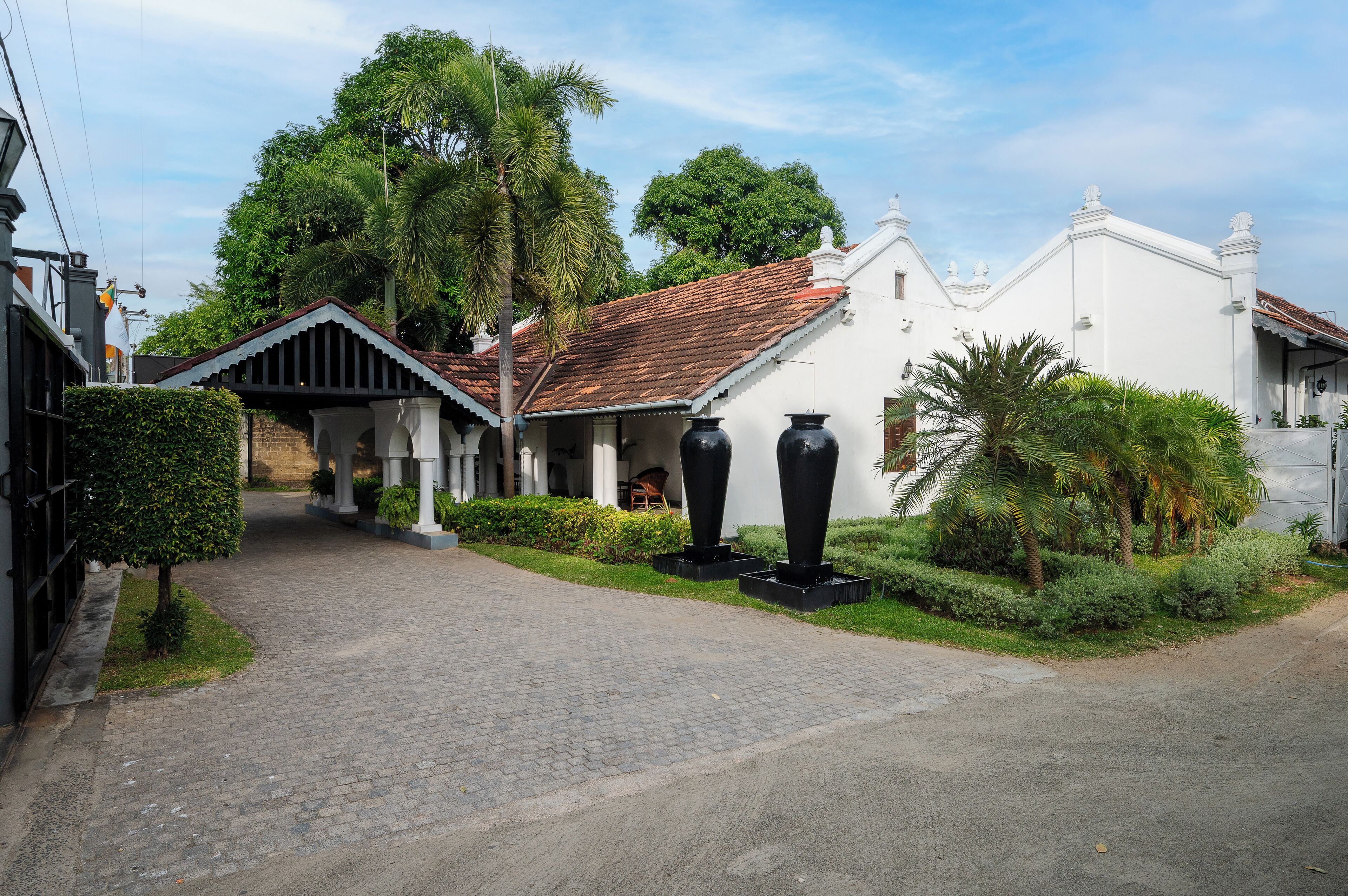A pathway leading to a building with a tiled roof and white walls, surrounded by greenery and palm trees.