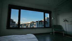 A hotel room with a large window, a bed, a chair, and a floor lamp. - Morning Dew Guesthouse (Busan)